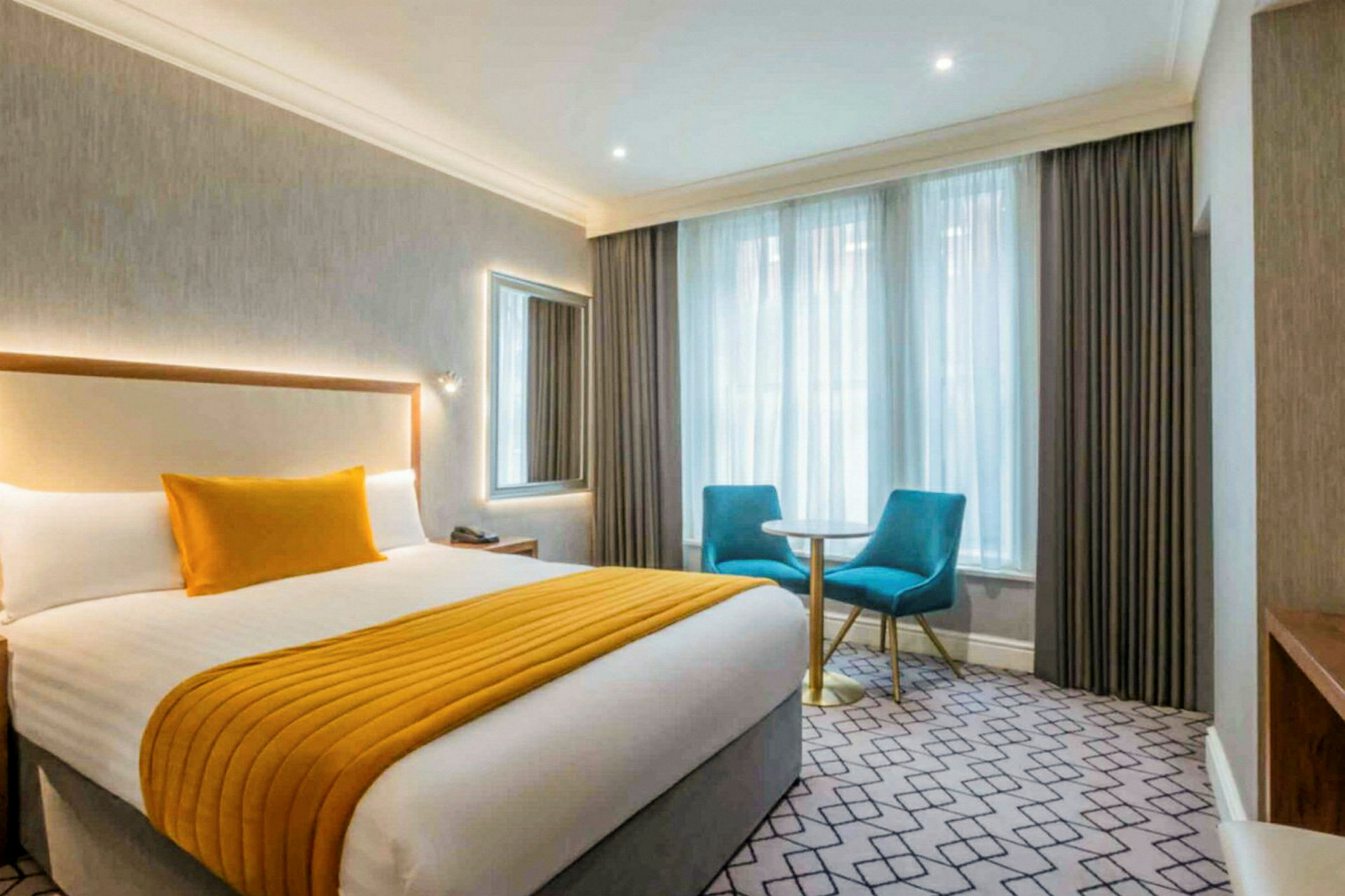 Hotelværelse med en dobbeltseng, to blå stole og et lille bord ved vinduet. (AI-genereret tekst)