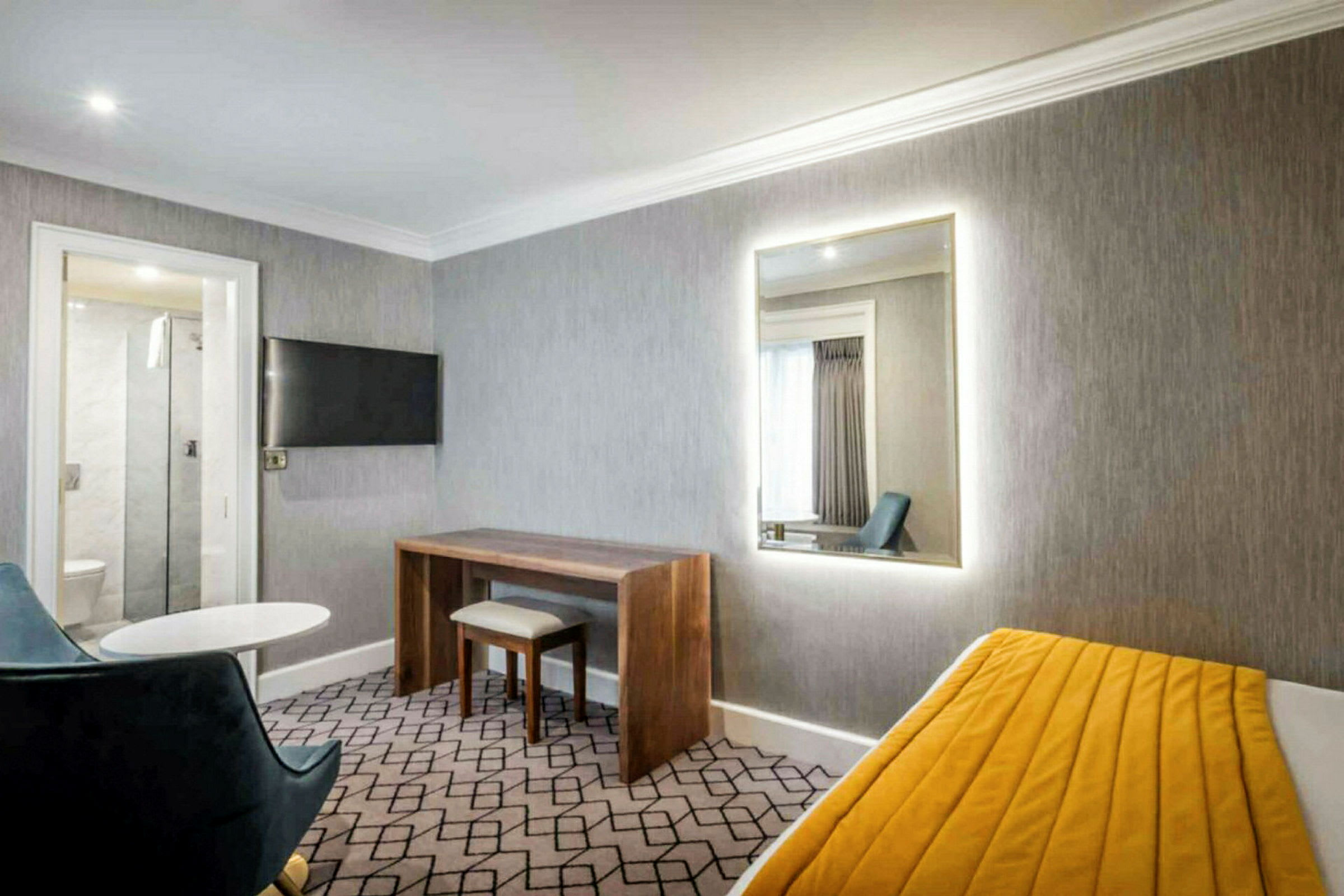Et hotelværelse med grå vægge, et spejl, et skrivebord, en stol og en seng med gult tæppe. (AI-genereret tekst)