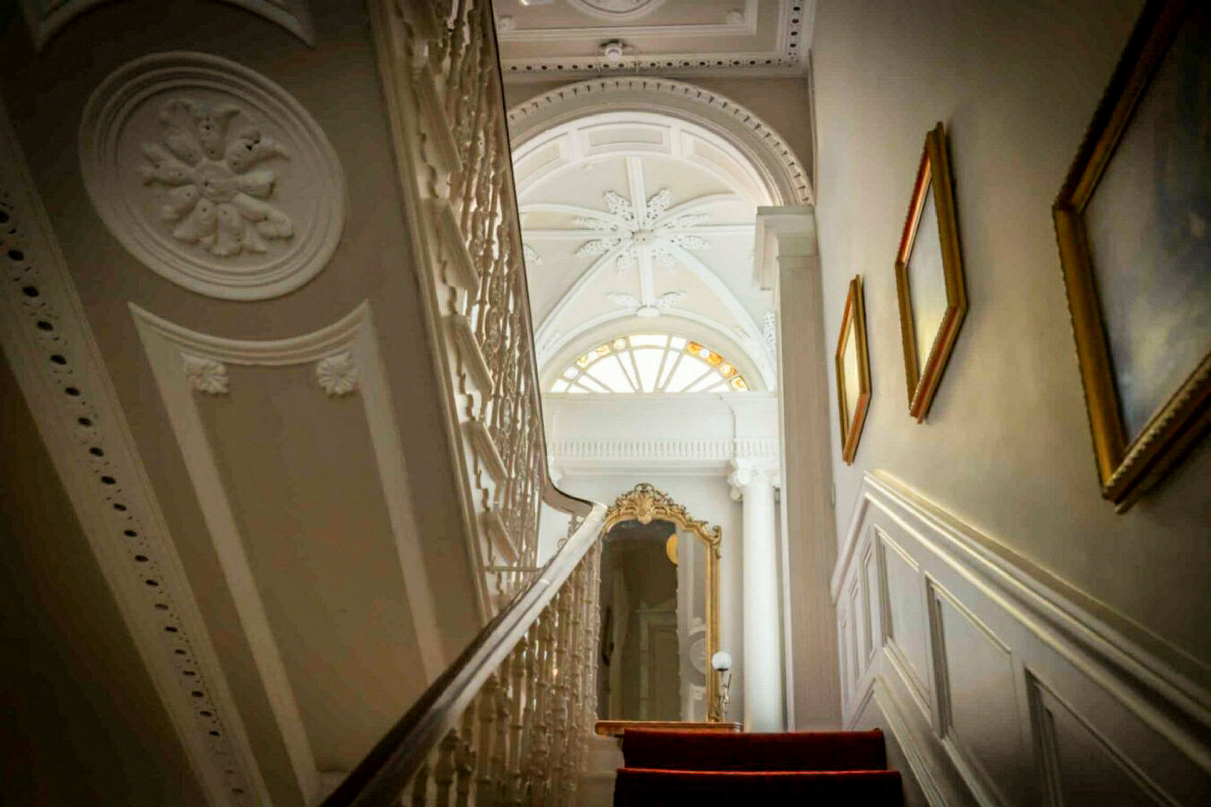 En trappe med rødt tæppe fører op til et rum med elegant loft og en stor vinduesformation. (AI-genereret tekst)