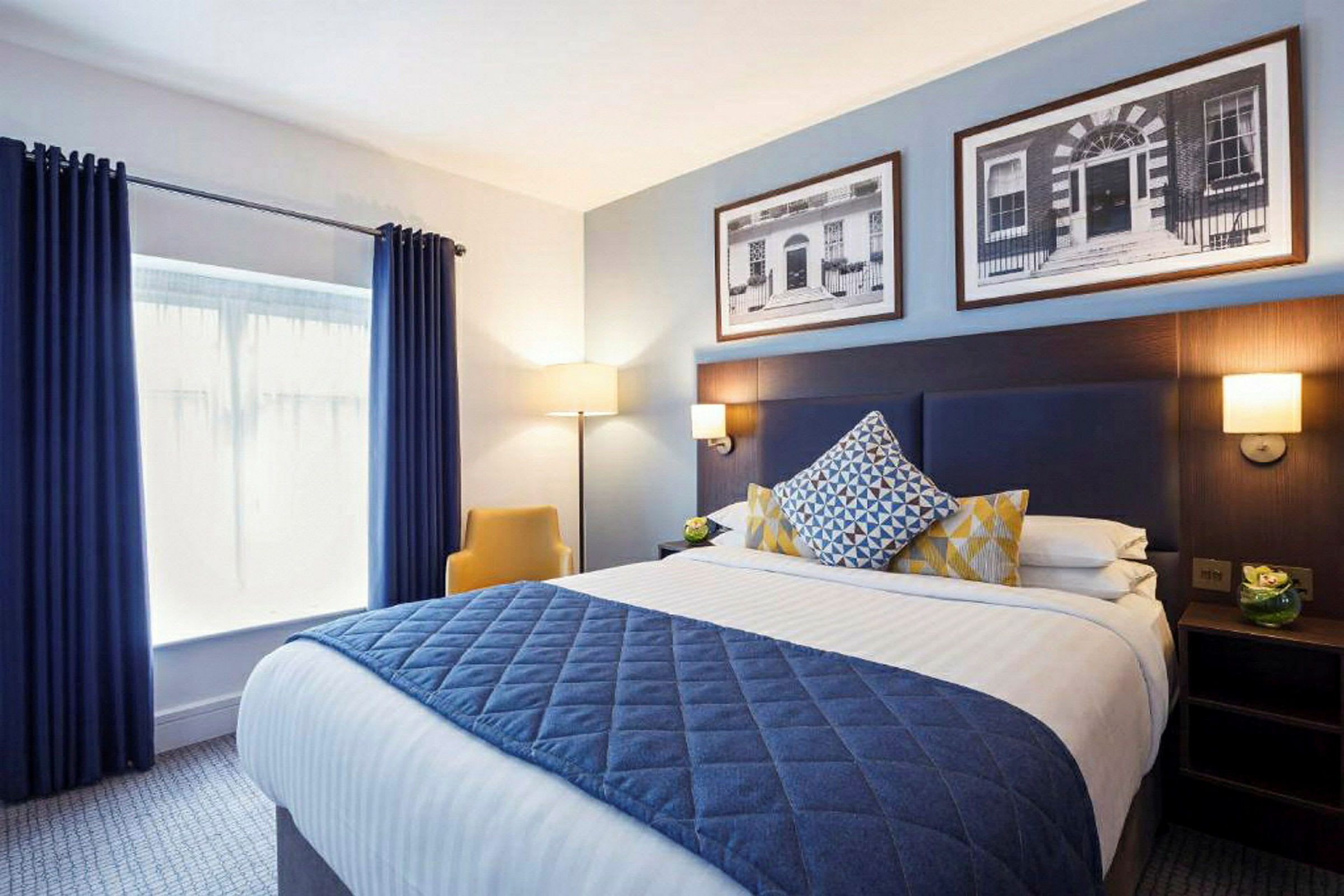 Moderne indrettet hotelværelse med en dobbeltseng, dekorerede puder og et tæppe i blå tone. (AI-genereret tekst)