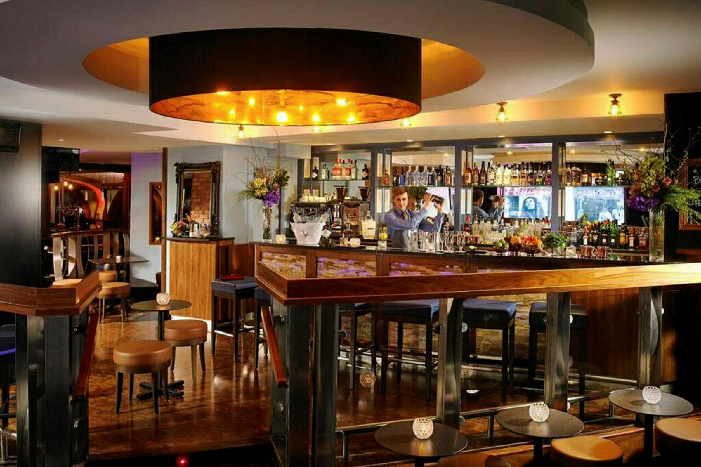 En elegant bar med træinteriør, bardisk og stole samt en bartender, der arbejder. (AI-genereret tekst)