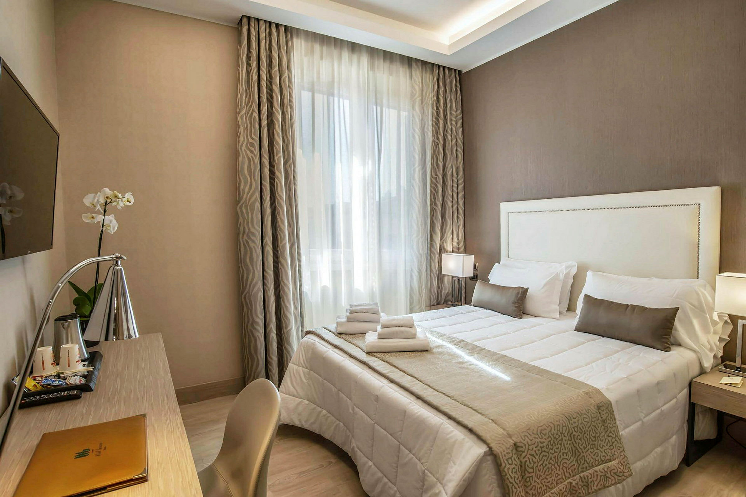 Et hotelværelse med en dobbeltseng med hvidt og beige sengetøj, et skrivebord med en lampe og et vægmonteret TV. (AI-genereret tekst)