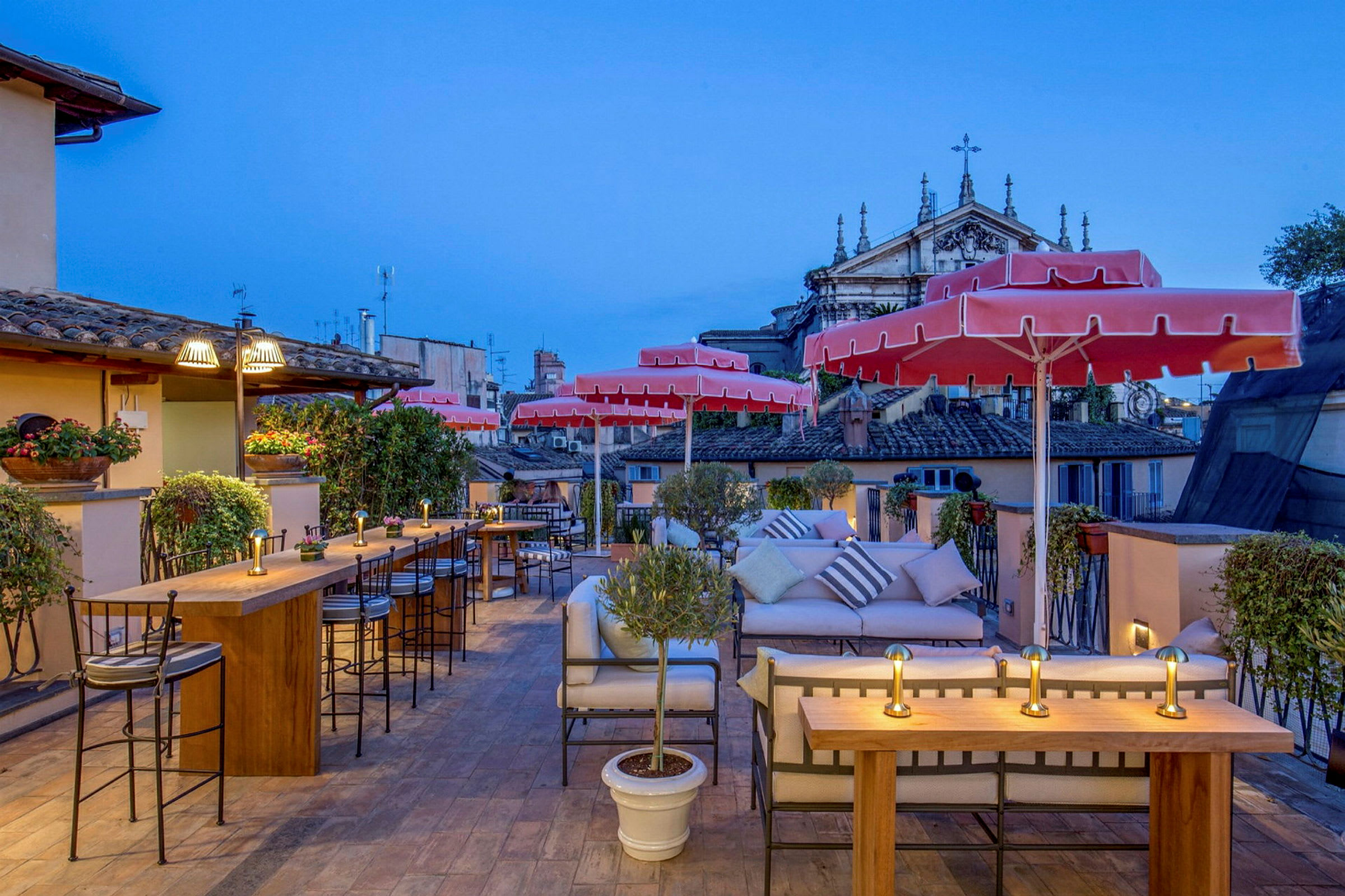 En terrasse med borde og stole, røde parasoller, grønne planter og udsigt over byens bygninger. (AI-genereret tekst)