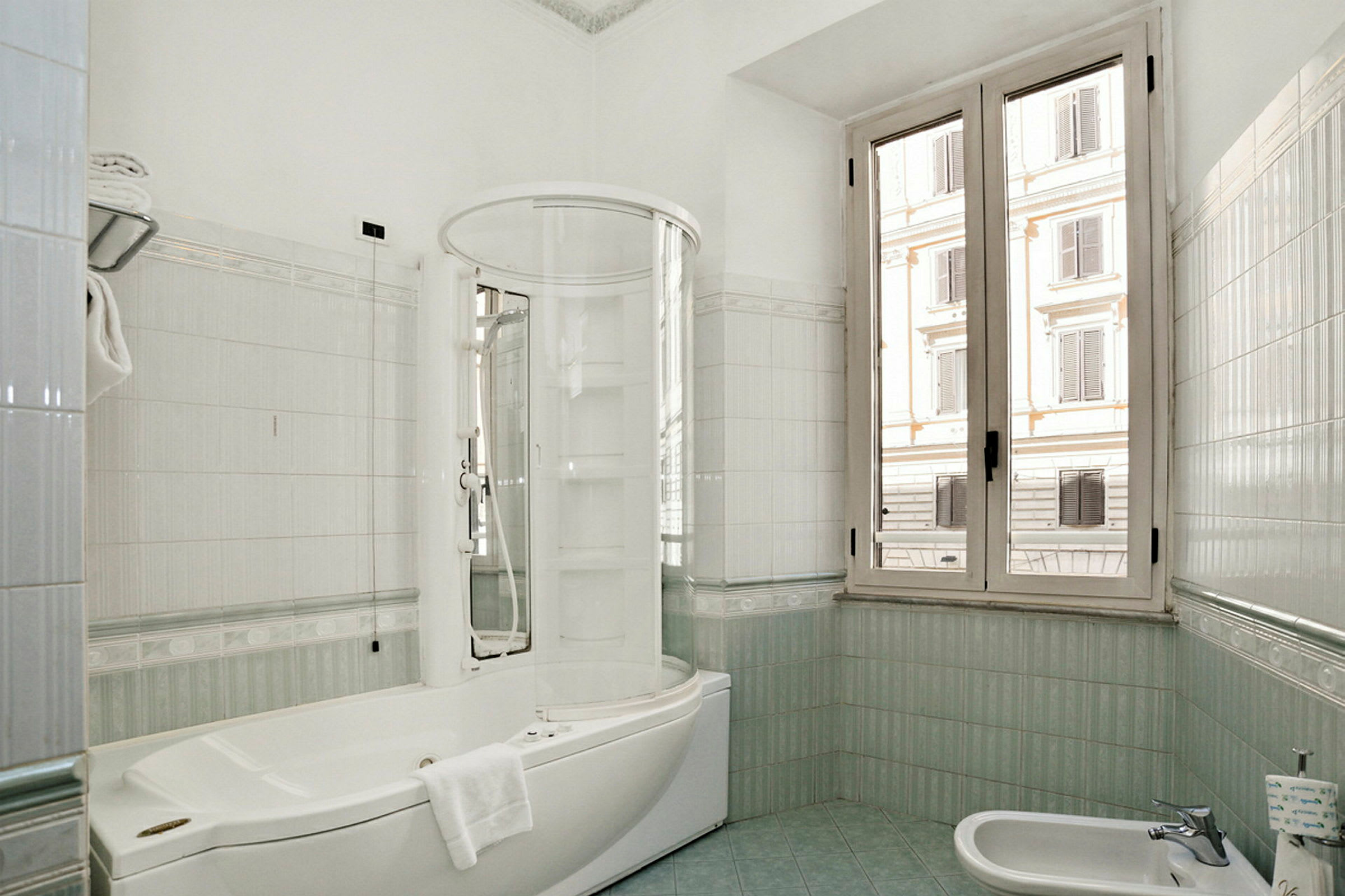 Et lyst badeværelse med stort vindue, hvidt badekar med bruser og lille bidet. (AI-genereret tekst)
