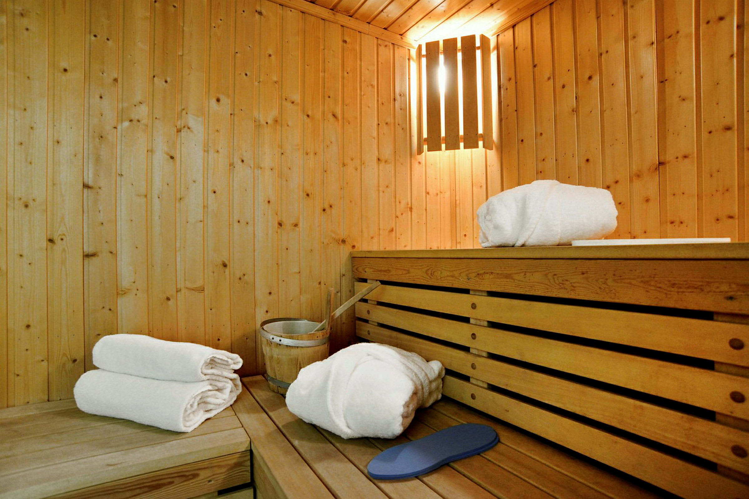 En sauna med træpaneler, oplyst af en pære. På træbænken ligger hvide håndklæder og et saunaaggregat. (AI-genereret tekst)