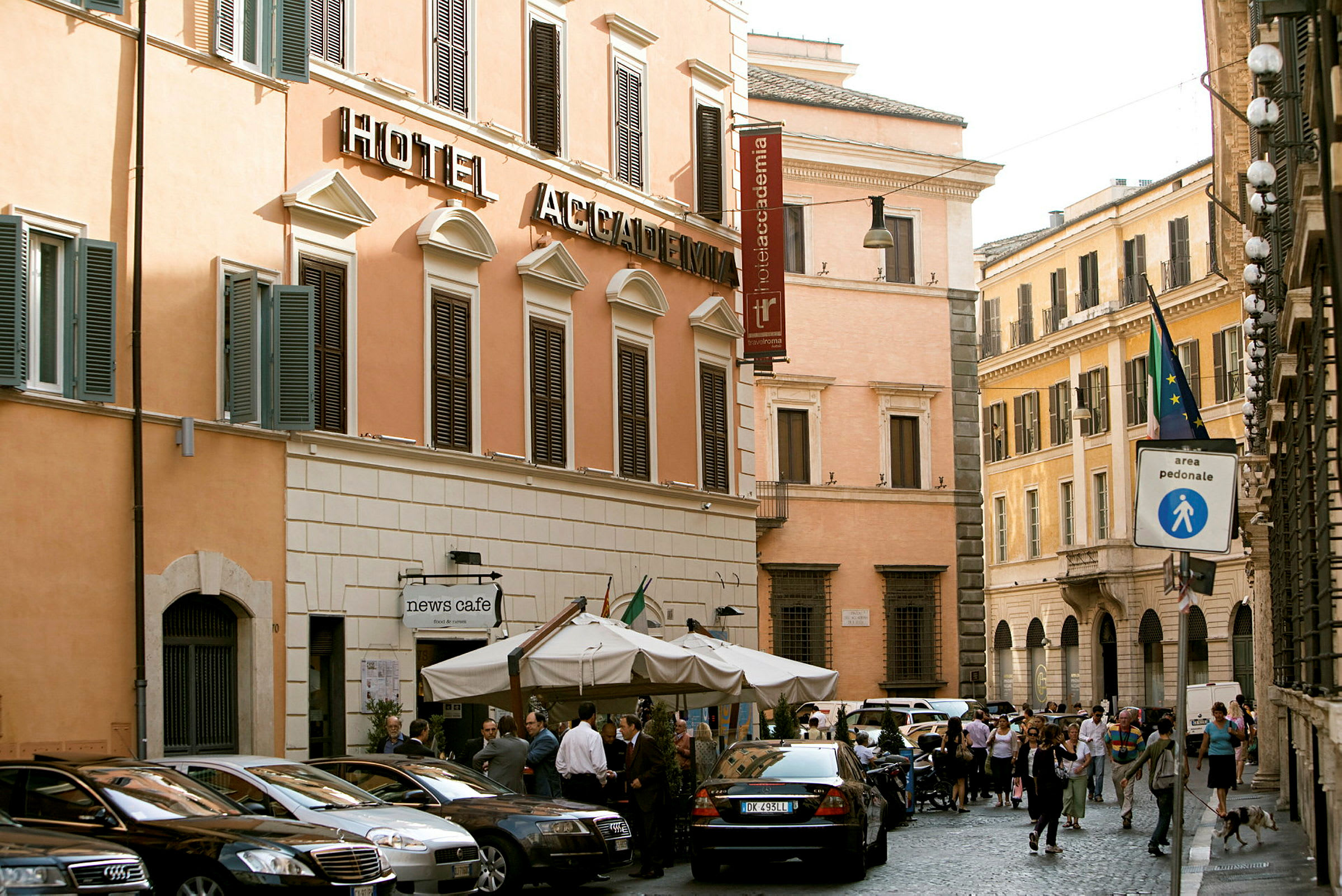 En gadescene med en hotelbygning med beige facade og mørke skodder. Foran bygningen står flere parkede biler og en café med parasoller. (AI-genereret tekst)