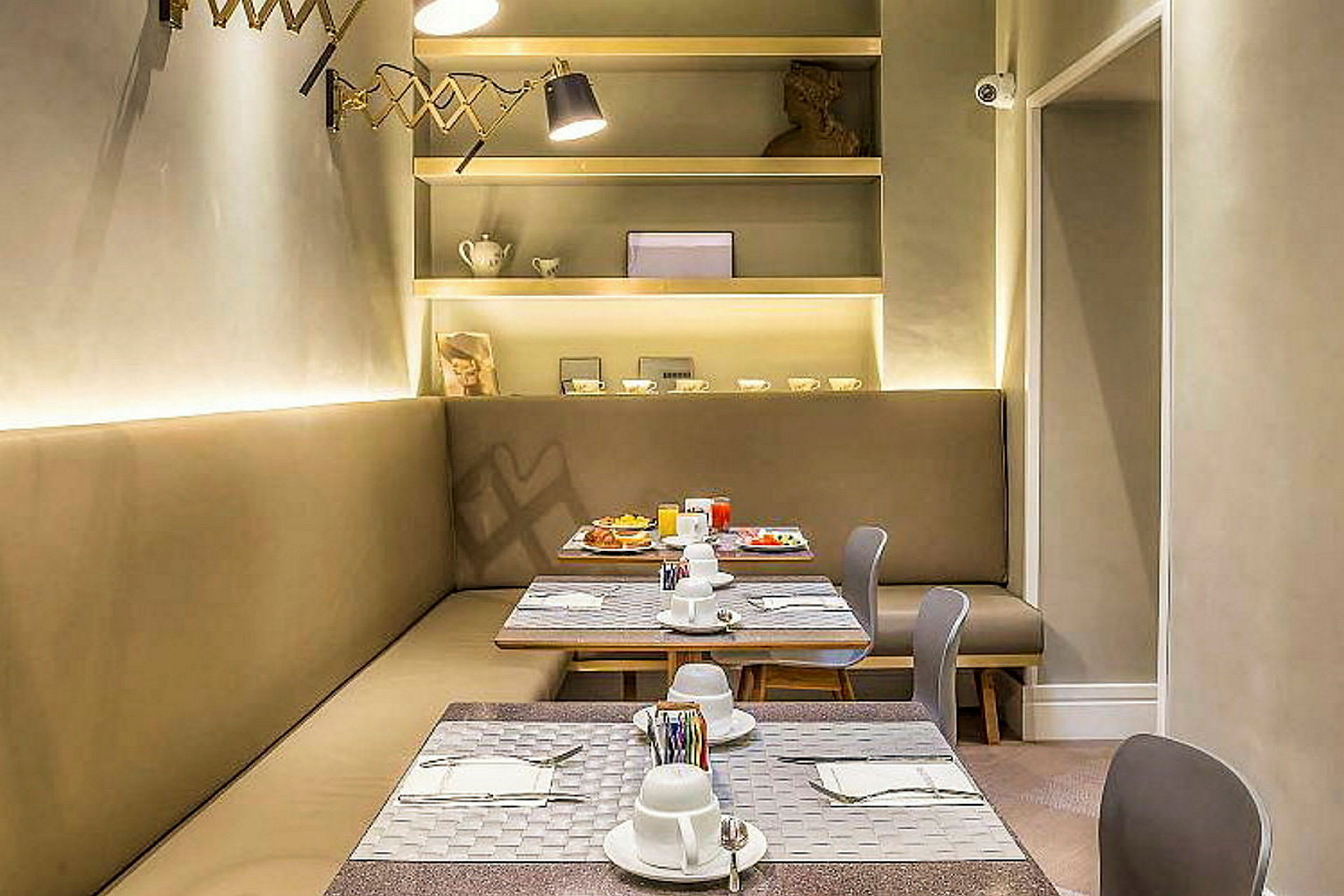 Et moderne møbleret restaurantområde med borde, stole og en siddebænk. Bordet er dækket med porcelæn og bestik. På væggen er der hylder med dekorative genstande. (AI-genereret tekst)