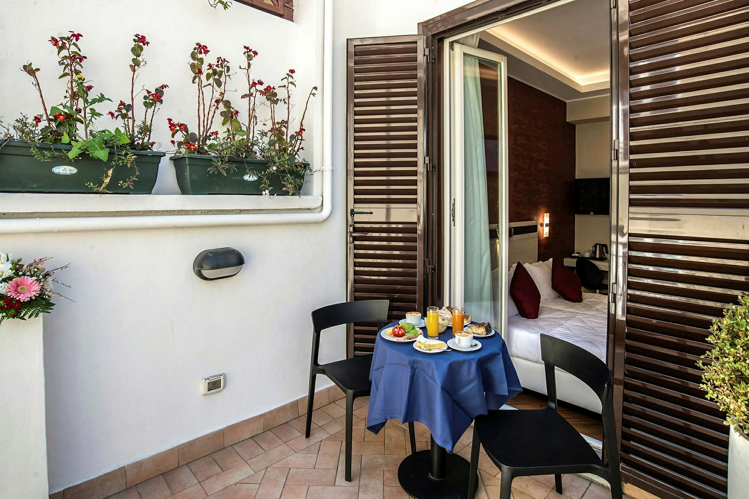En balkon med et lille bord dækket til morgenmad og to stole. Bordet er dækket med en blå dug og fører ind til et soveværelse. (AI-genereret tekst)