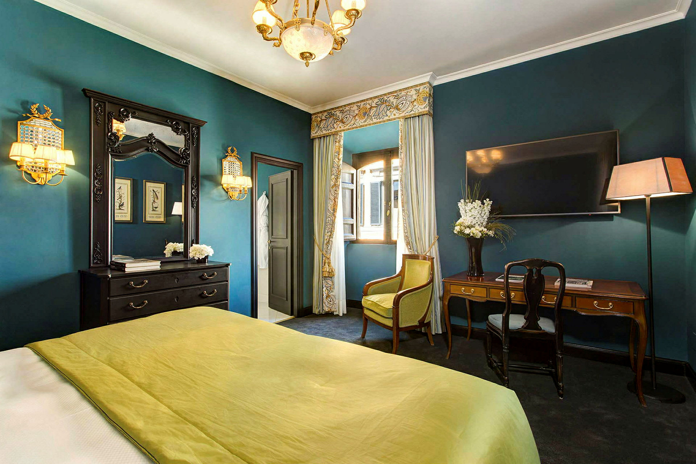 Et elegant indrettet hotelværelse med grønt sengetæppe, skrivebord, spejl og gardiner foran et åbent vindue. (AI-genereret tekst)
