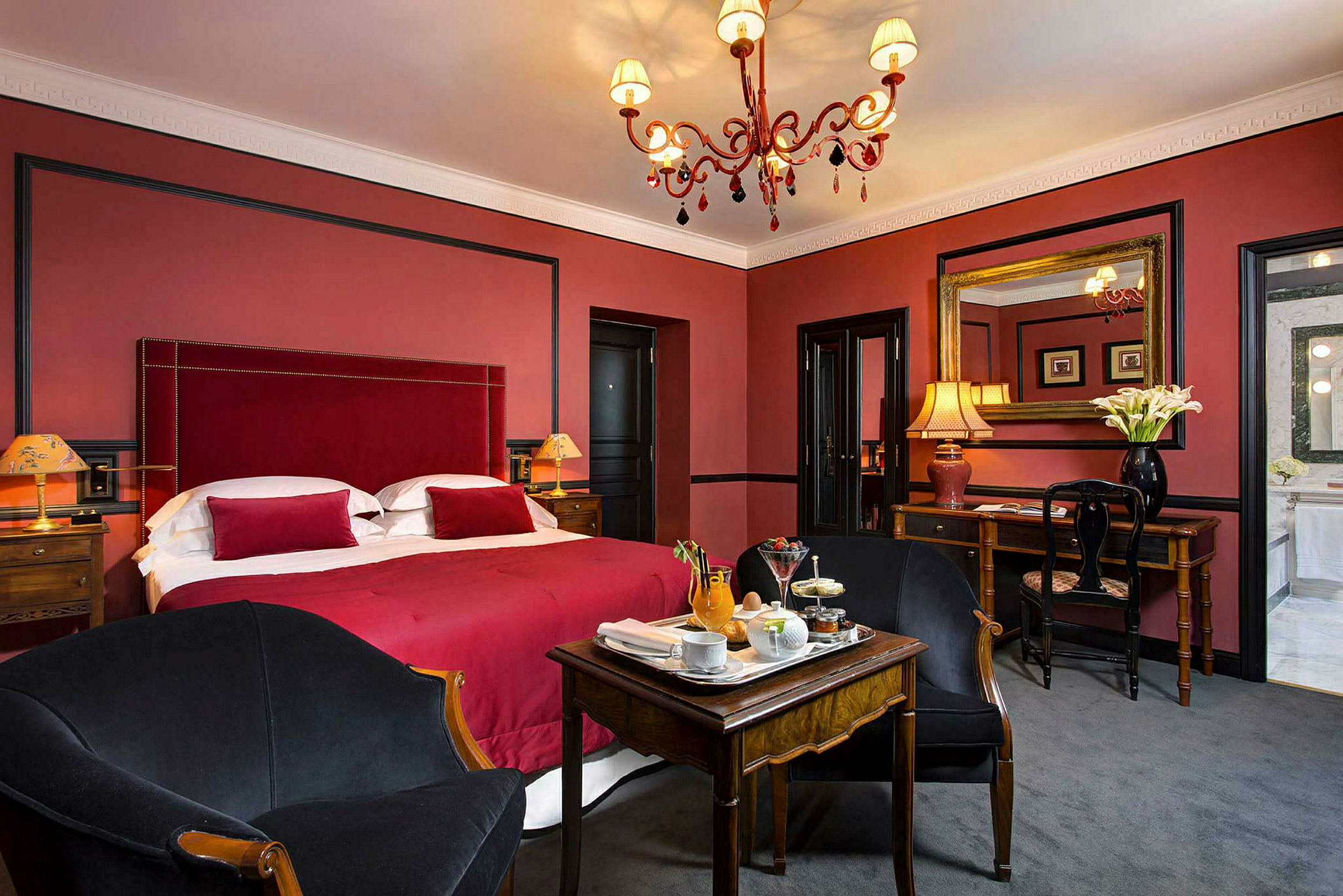 Et elegant hotelværelse med en rød sengegavl og sengetøj, en lysekrone og et siddeområde ved et lille træbord. (AI-genereret tekst)