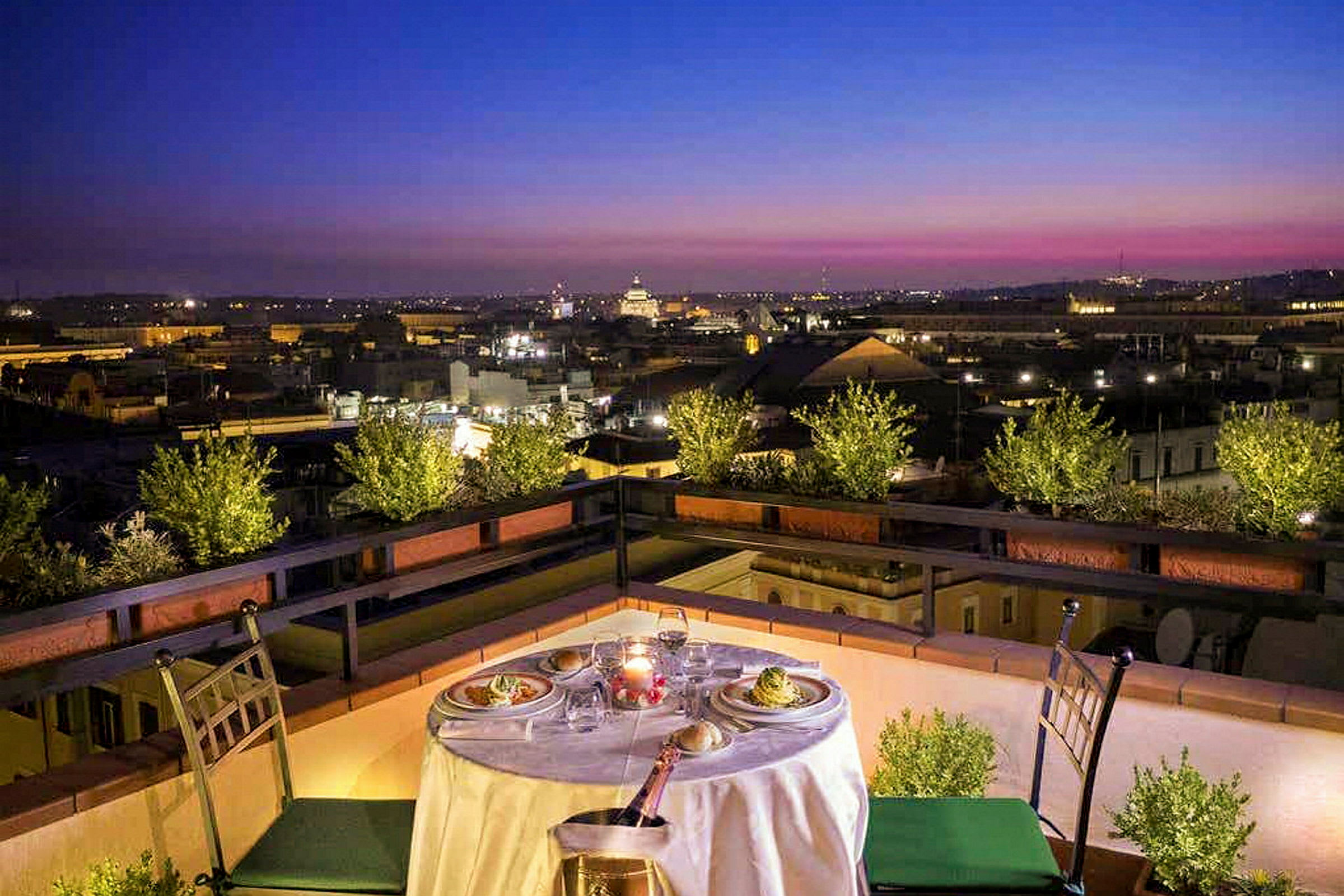 Et dækket bord med mad og drikke på en terrasse med udsigt over en by ved solnedgang. (AI-genereret tekst)