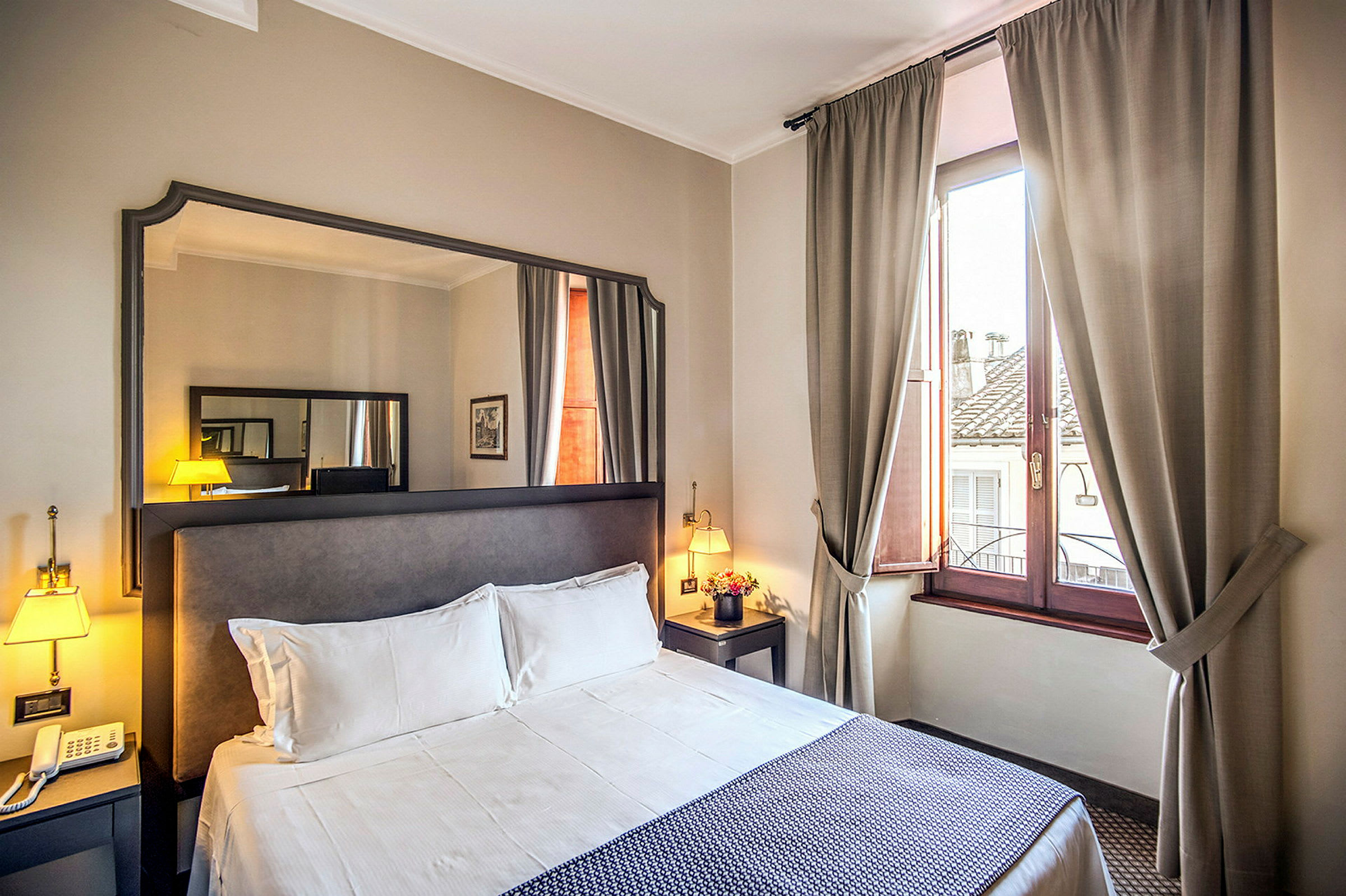 Hotelværelse med dobbeltseng, natbord, spejl og vindue med beige gardiner. (AI-genereret tekst)