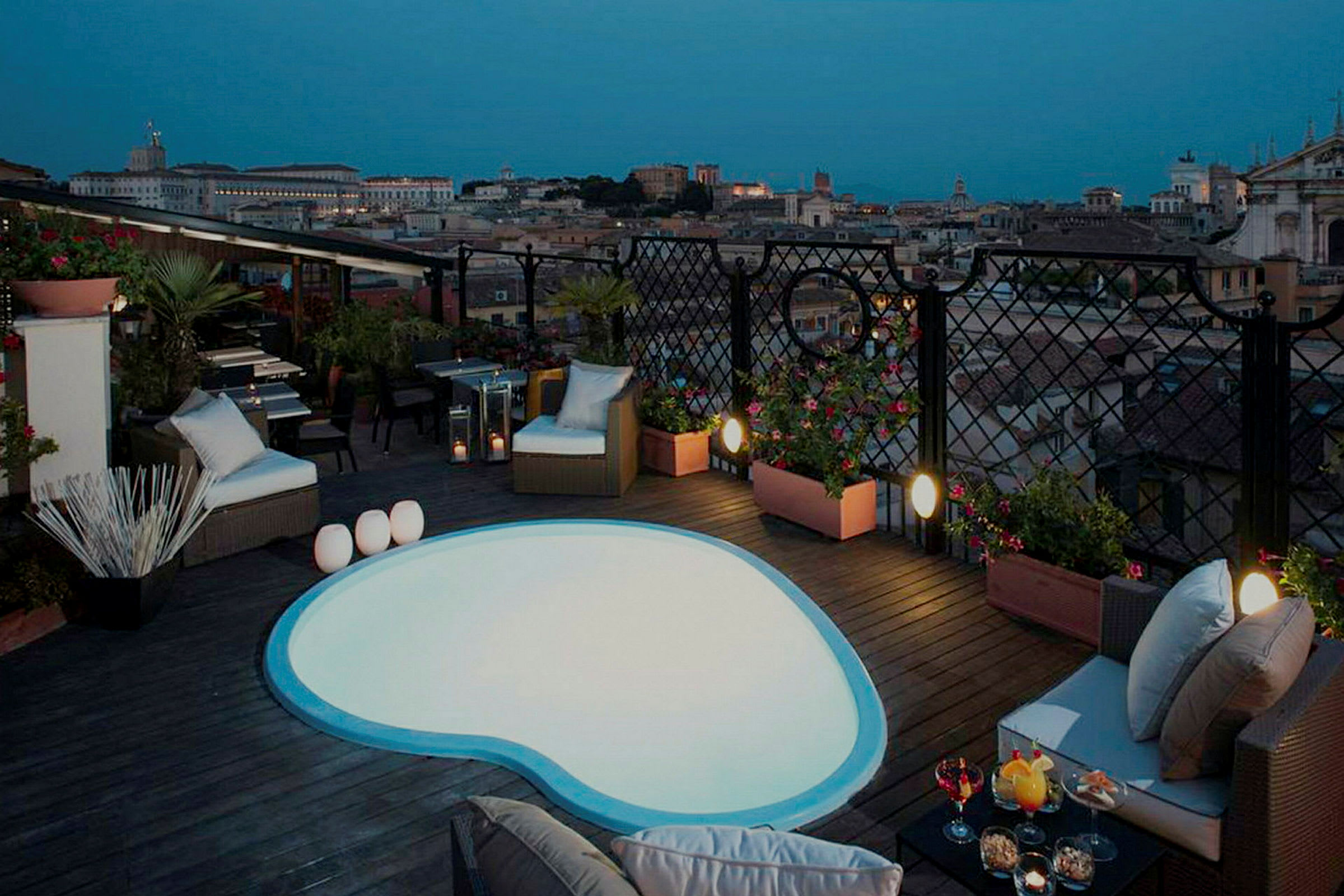 Billede af en tagterrasse med en lille pool og siddepladser samt udsigt over en by i skumringen. (AI-genereret tekst)