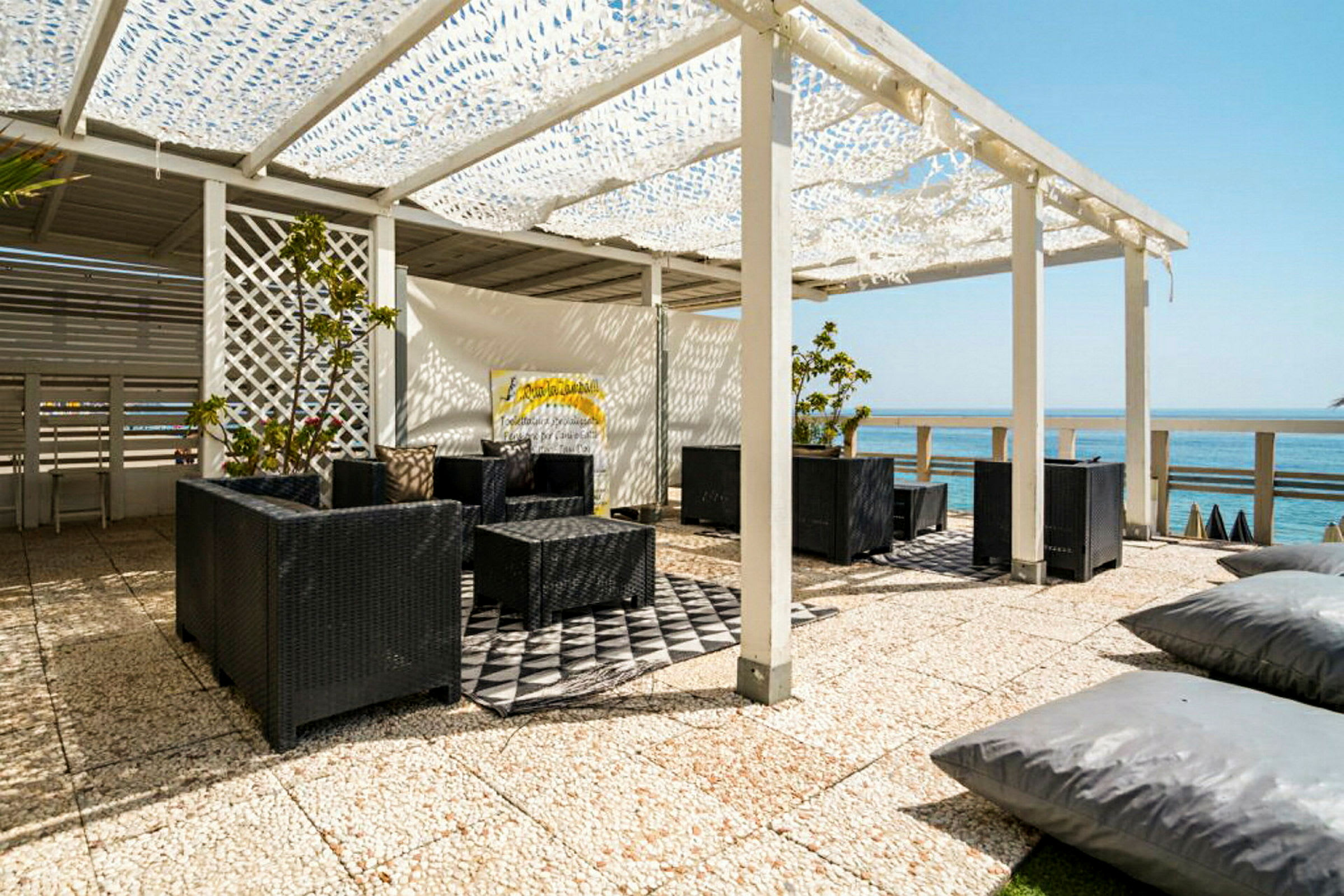 Udendørs siddeområde med sorte rottingmøbler under en hvid pergola med udsigt over havet. (AI-genereret tekst)