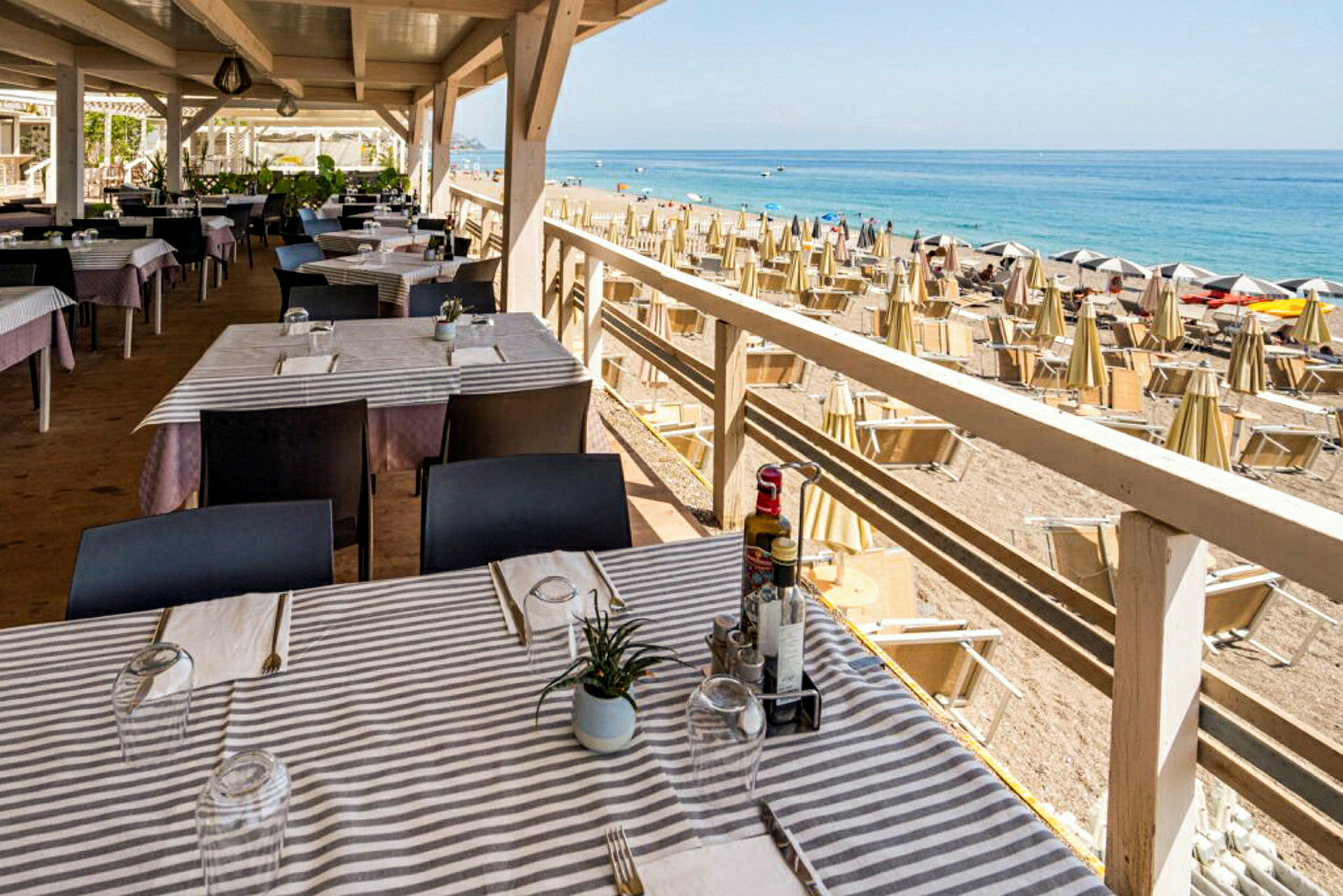 En restaurant med dækkede borde tæt på en strand med parasoller og liggestole. (AI-genereret tekst)
