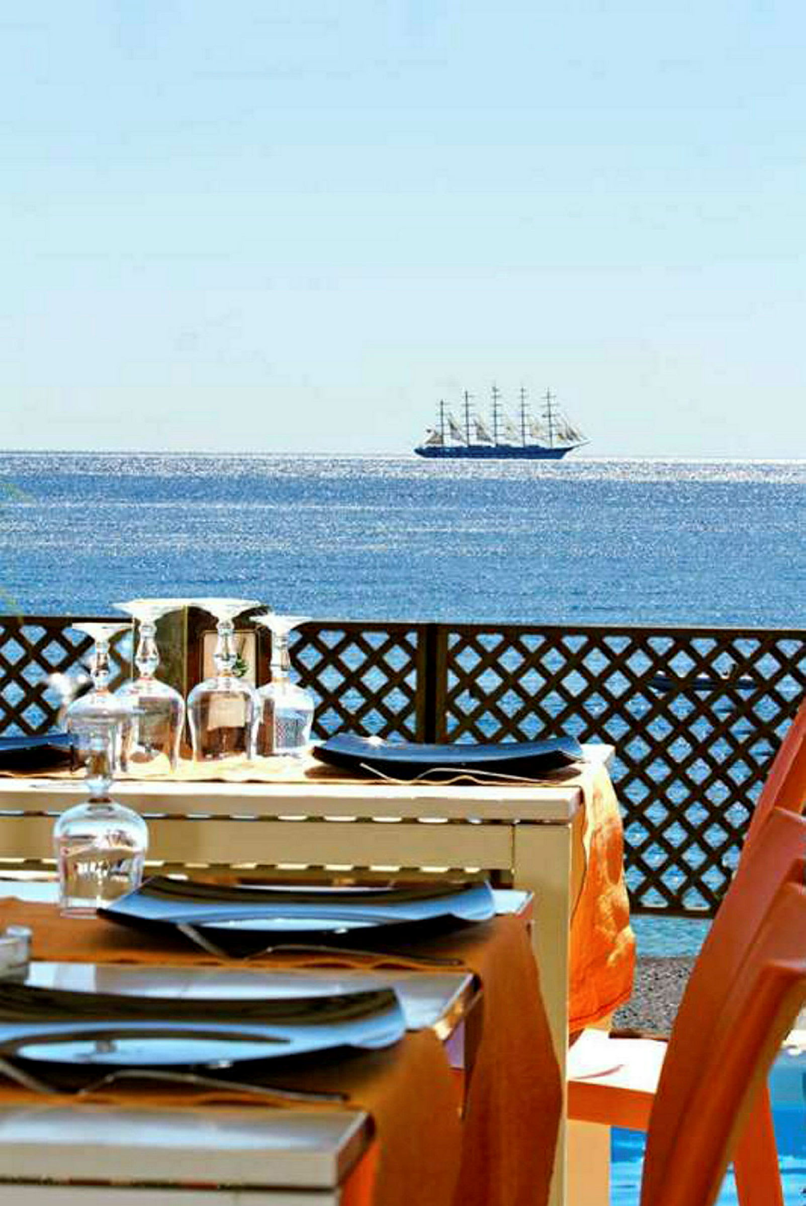Et dækket bord på en terrasse med udsigt over havet og et sejlskib i det fjerne. (AI-genereret tekst)