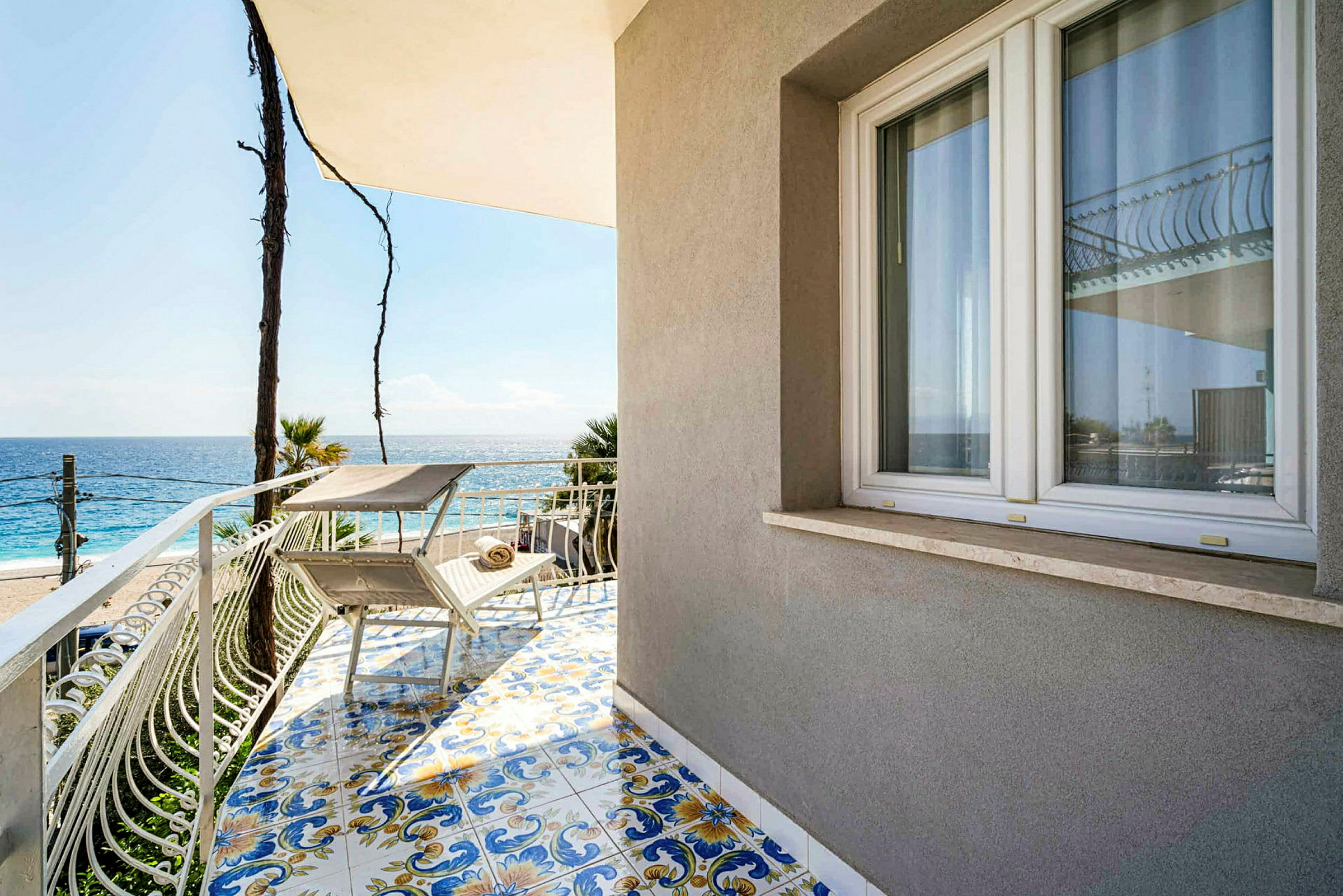 En balkon med dekorative fliser, to liggestole og udsigt over havet. (AI-genereret tekst)