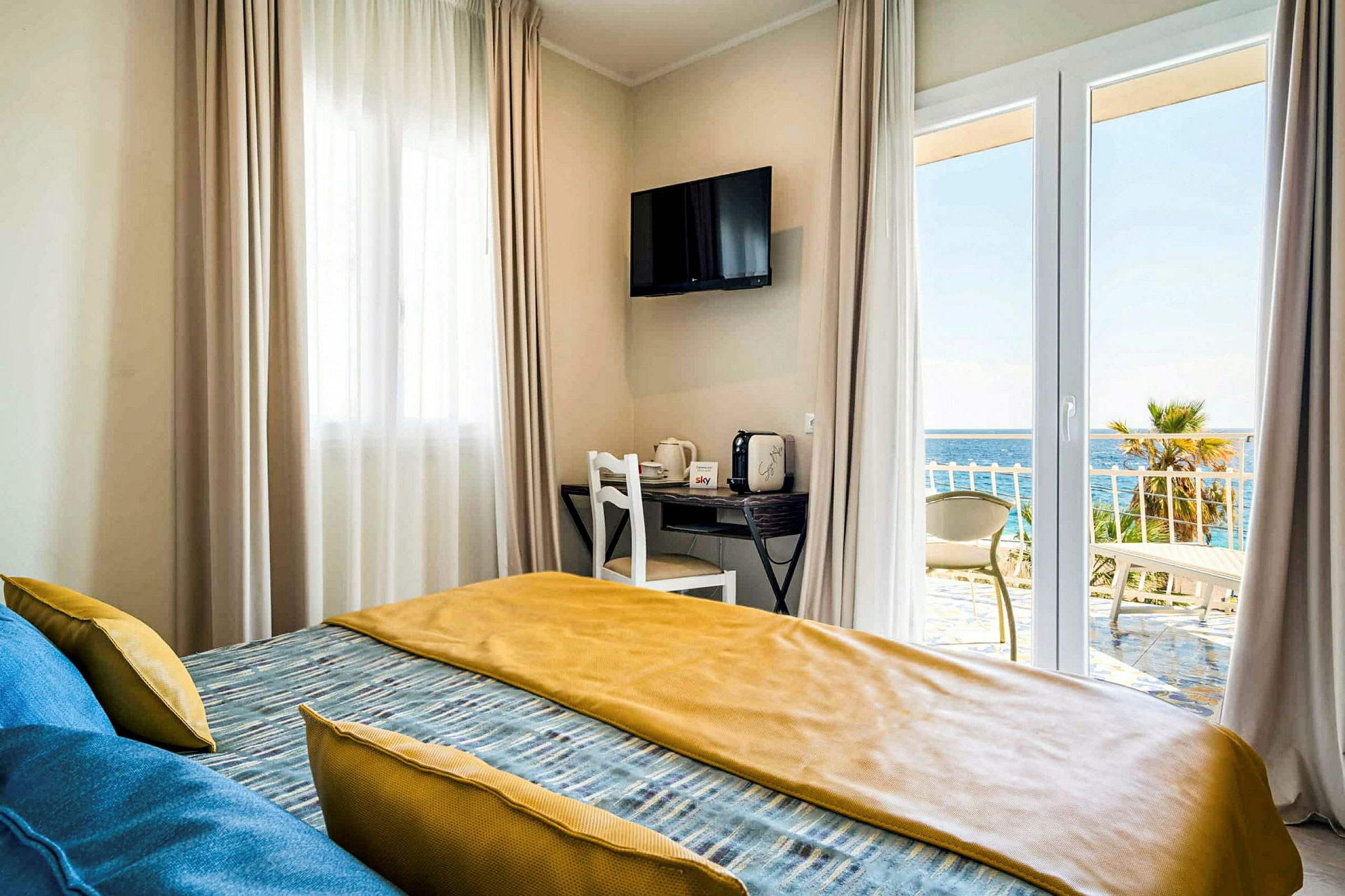 Et hotelværelse med en seng, vinduer med gardiner, et TV på væggen og udsigt over havet fra balkonen. (AI-genereret tekst)