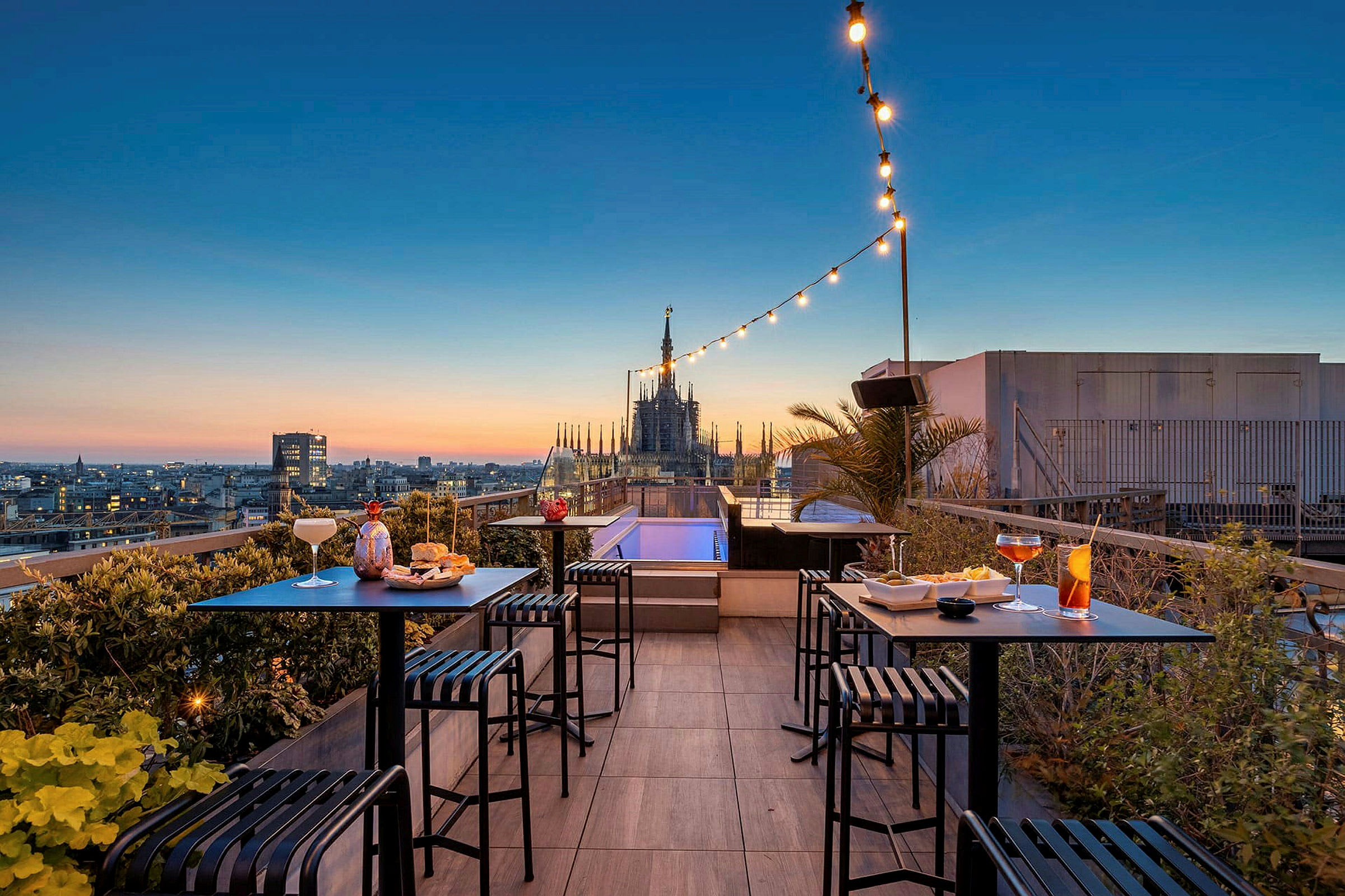 En tagterrasse med udsigt over en by ved solnedgang, hvor små borde og stole er placeret med drikkevarer og snacks. (AI-genereret tekst)