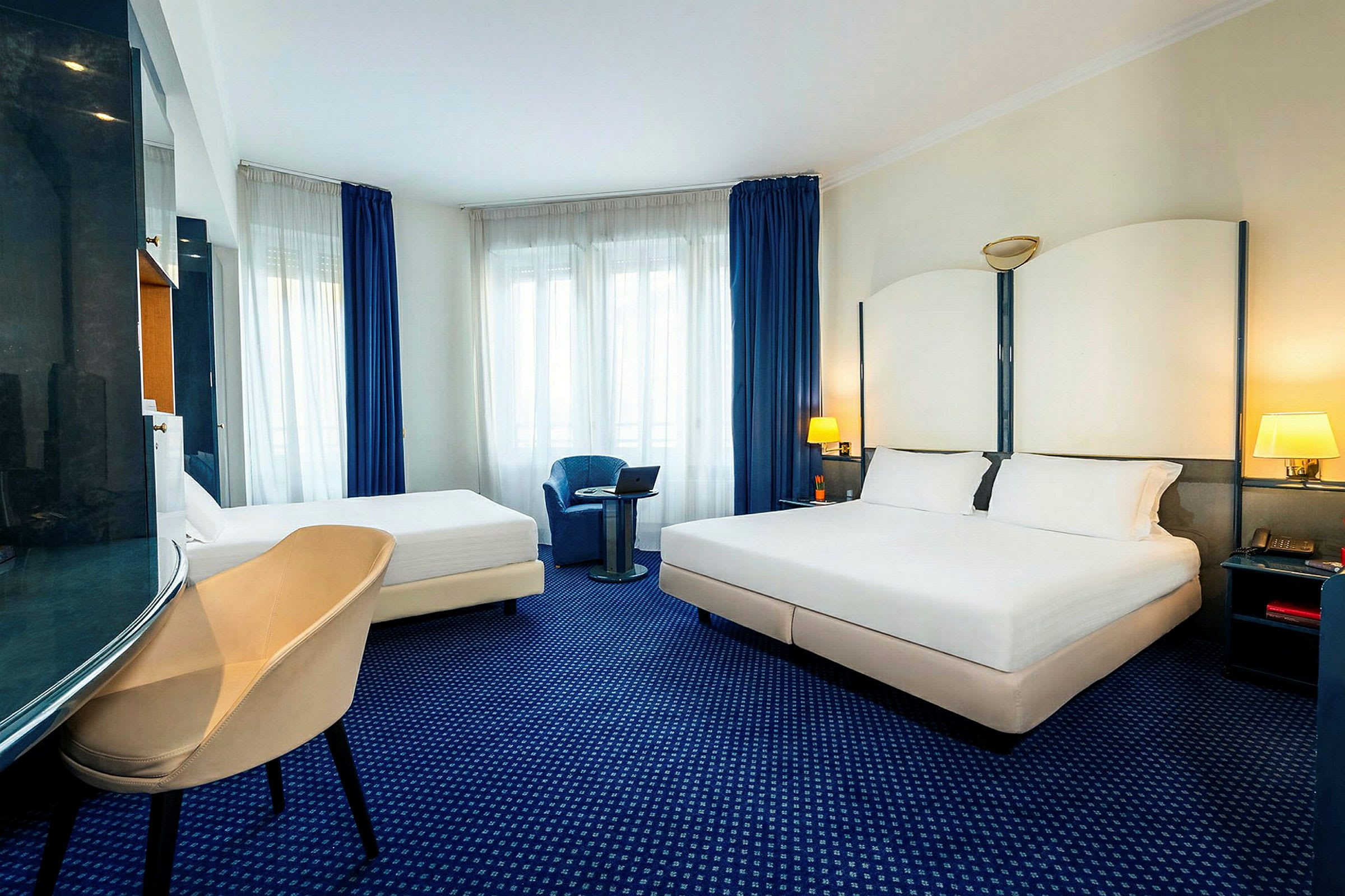 Hotelværelse med en dobbeltseng og en enkeltseng, lænestol og vindue med blå gardiner. (AI-genereret tekst)