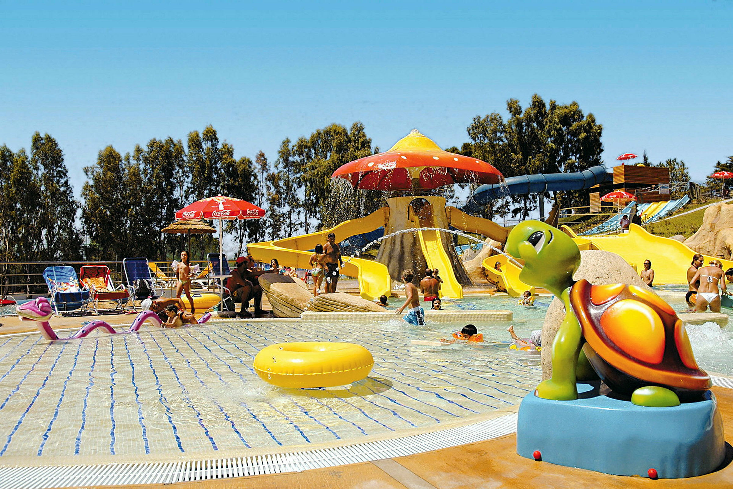 Billedet viser en vandpark med en lavvandet pool og flere vandrutsjebaner samt en skulptur af en farverig skildpadde. (AI-genereret tekst)