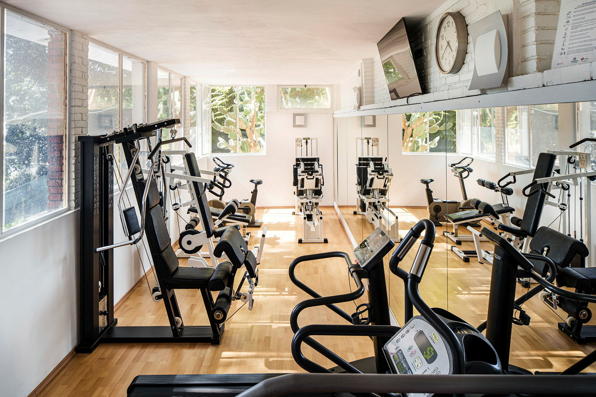 Et fitnesscenter med træningsmaskiner, herunder stationære cykler og løbebånd, i et lyst område. Væggene har store spejle og vinduer. (AI-genereret tekst)