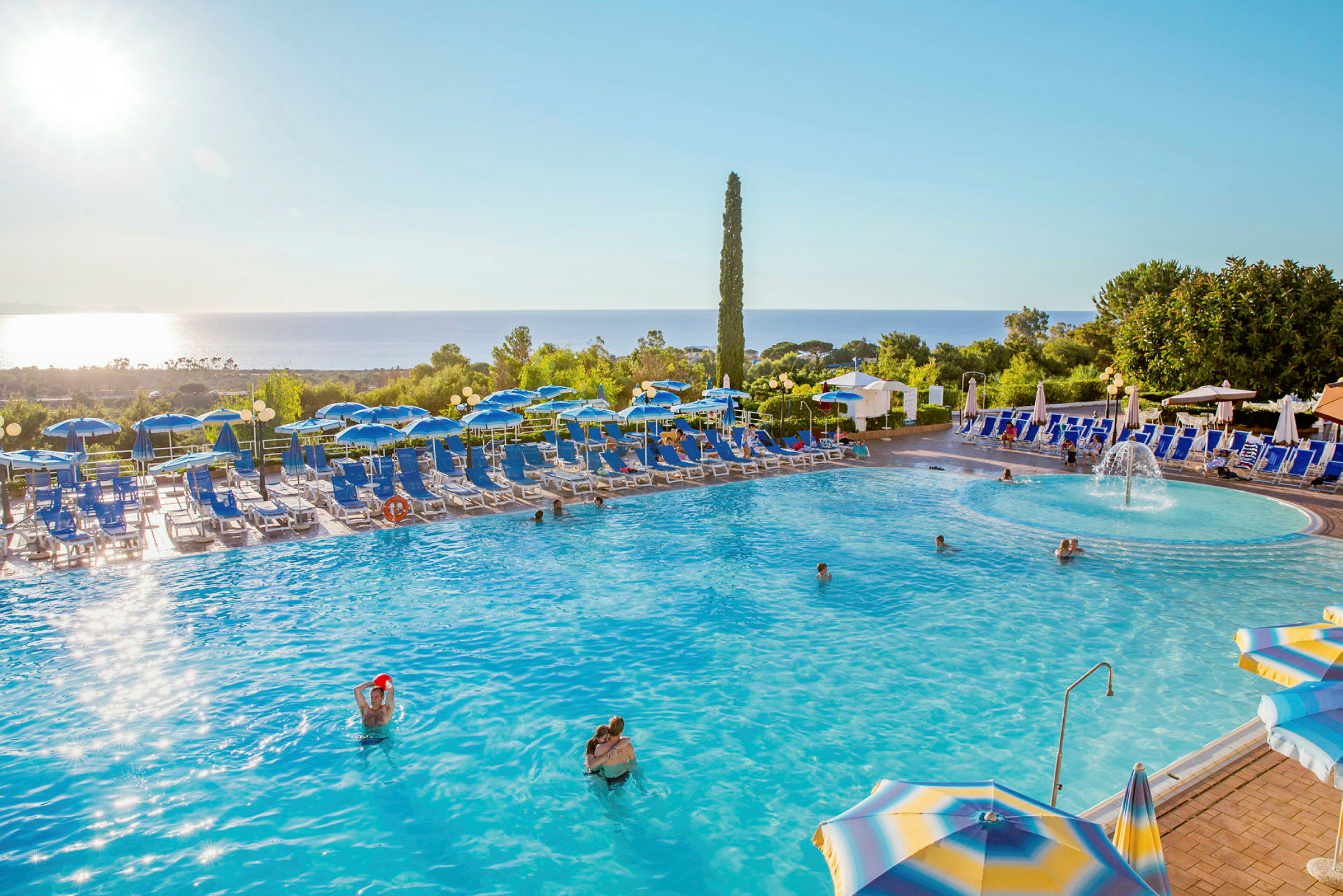 En swimmingpool med liggestole og parasoller omkring, omgivet af grønt og med havudsigt. (AI-genereret tekst)
