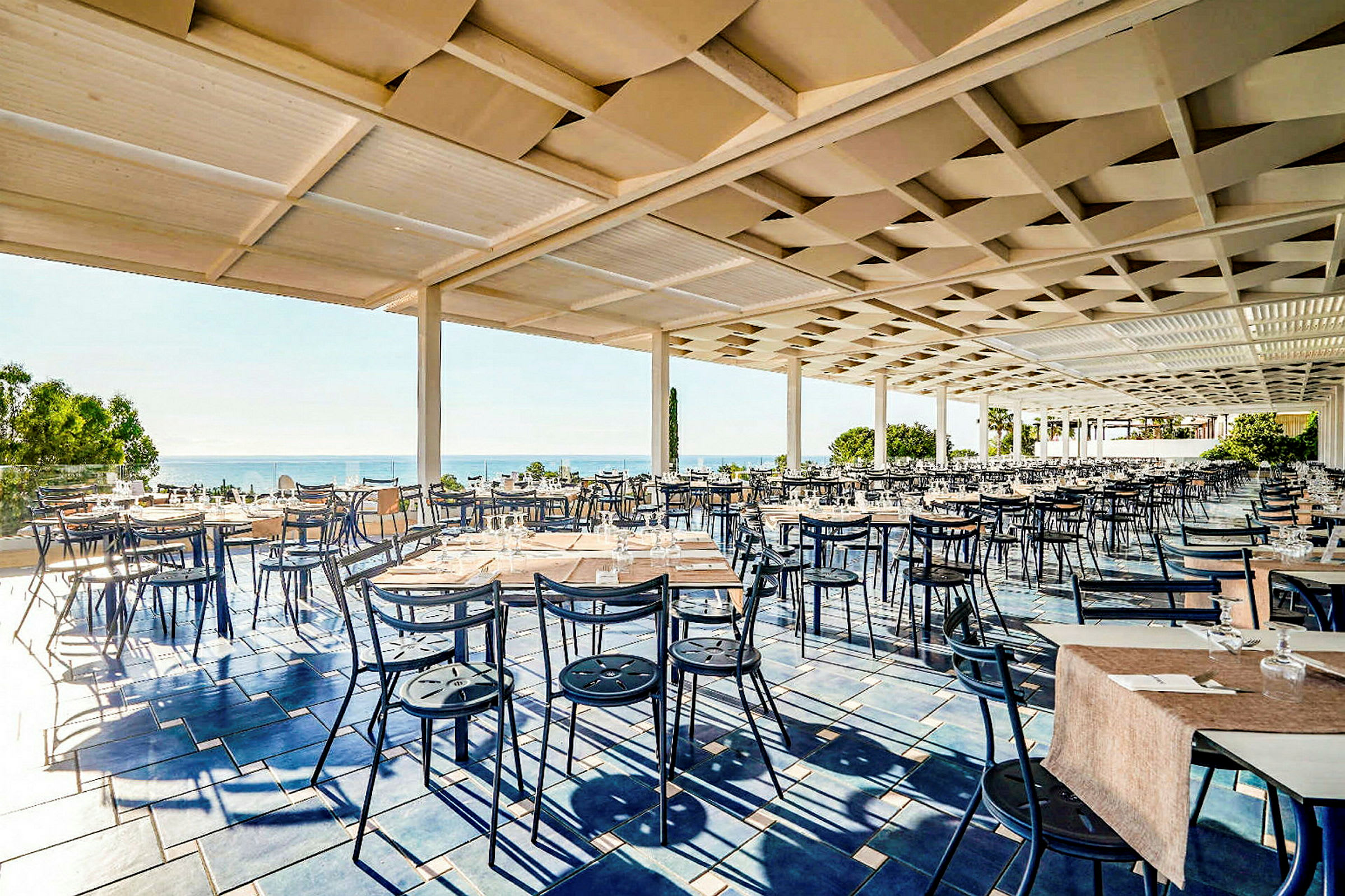 Udendørs restaurant med mange borde og stole, beliggende tæt på havet. (AI-genereret tekst)