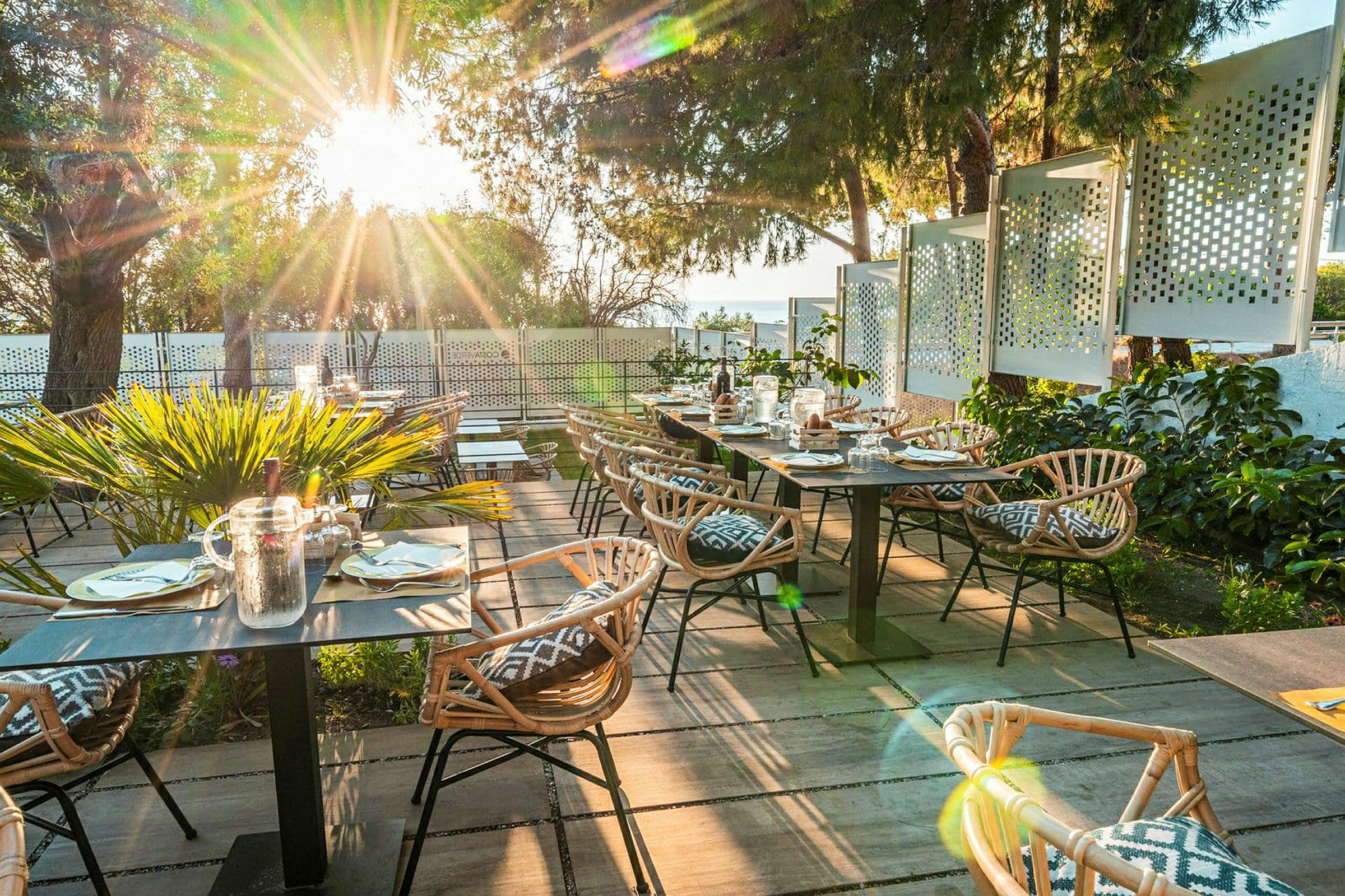 Udendørs restaurant med træborde og rottingstole samt solstråler gennem træerne. (AI-genereret tekst)