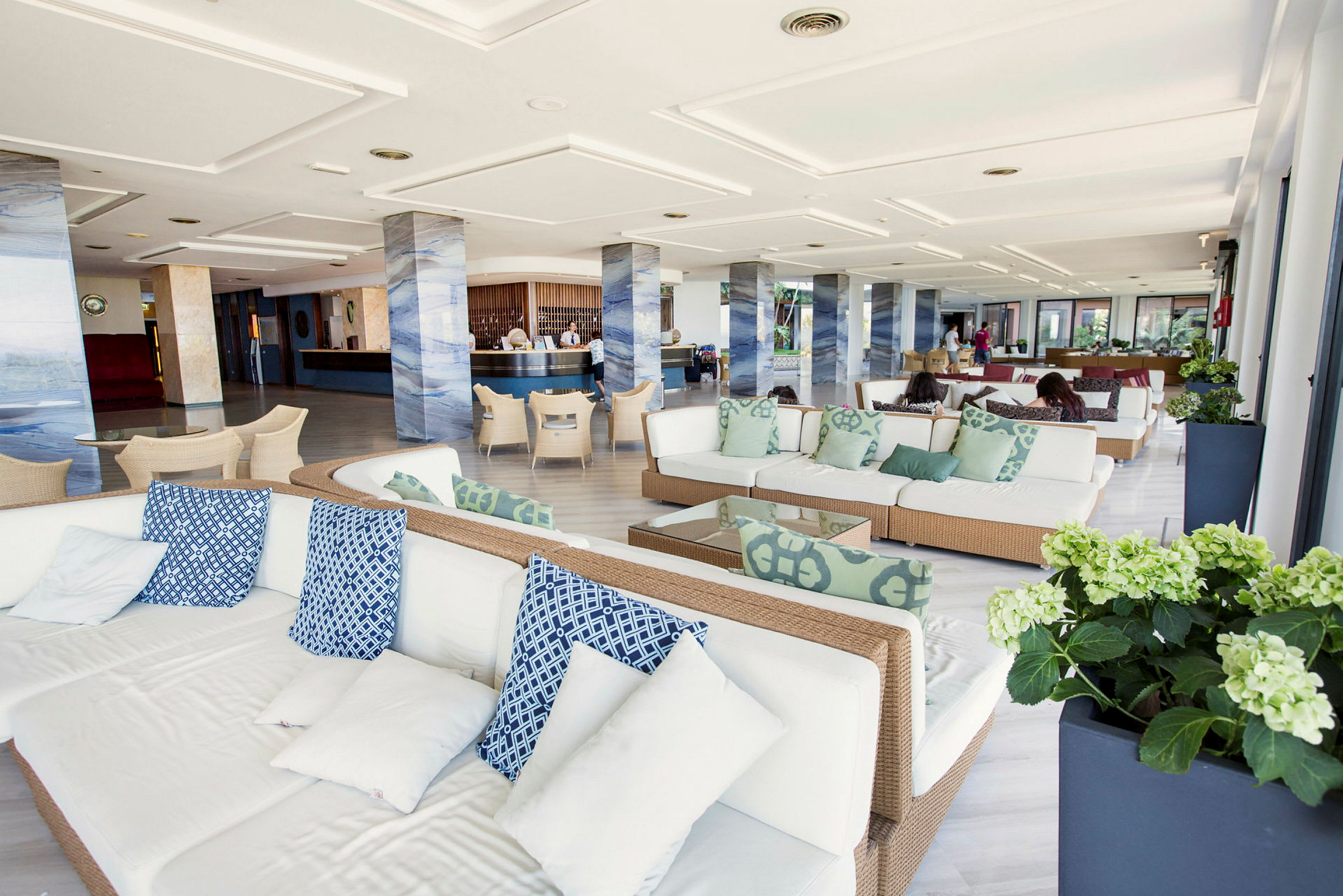 Et moderne lobbyområde med hvide sofaer og blå samt grønne puder. Området har søjler i et marmoreret design og et receptionsområde i baggrunden. (AI-genereret tekst)