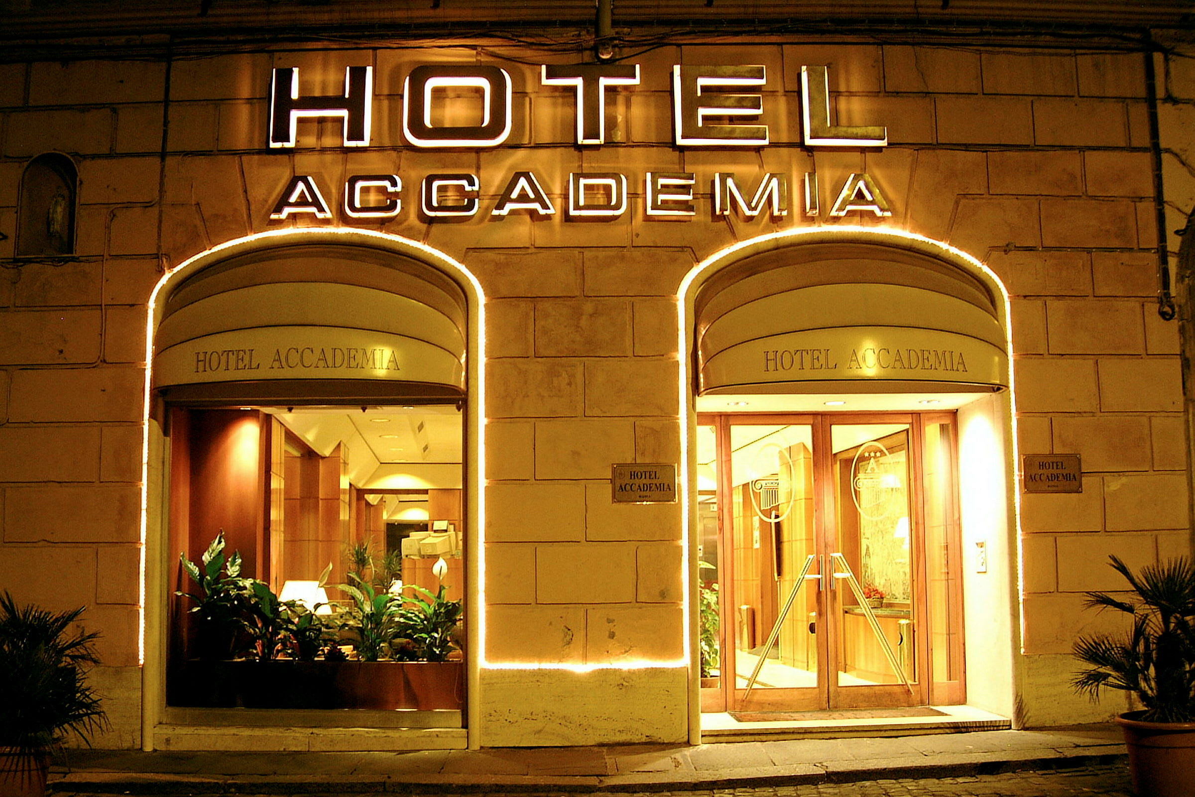 Hotel Accademias oplyste indgang med glasdøre og planter. (AI-genereret tekst)