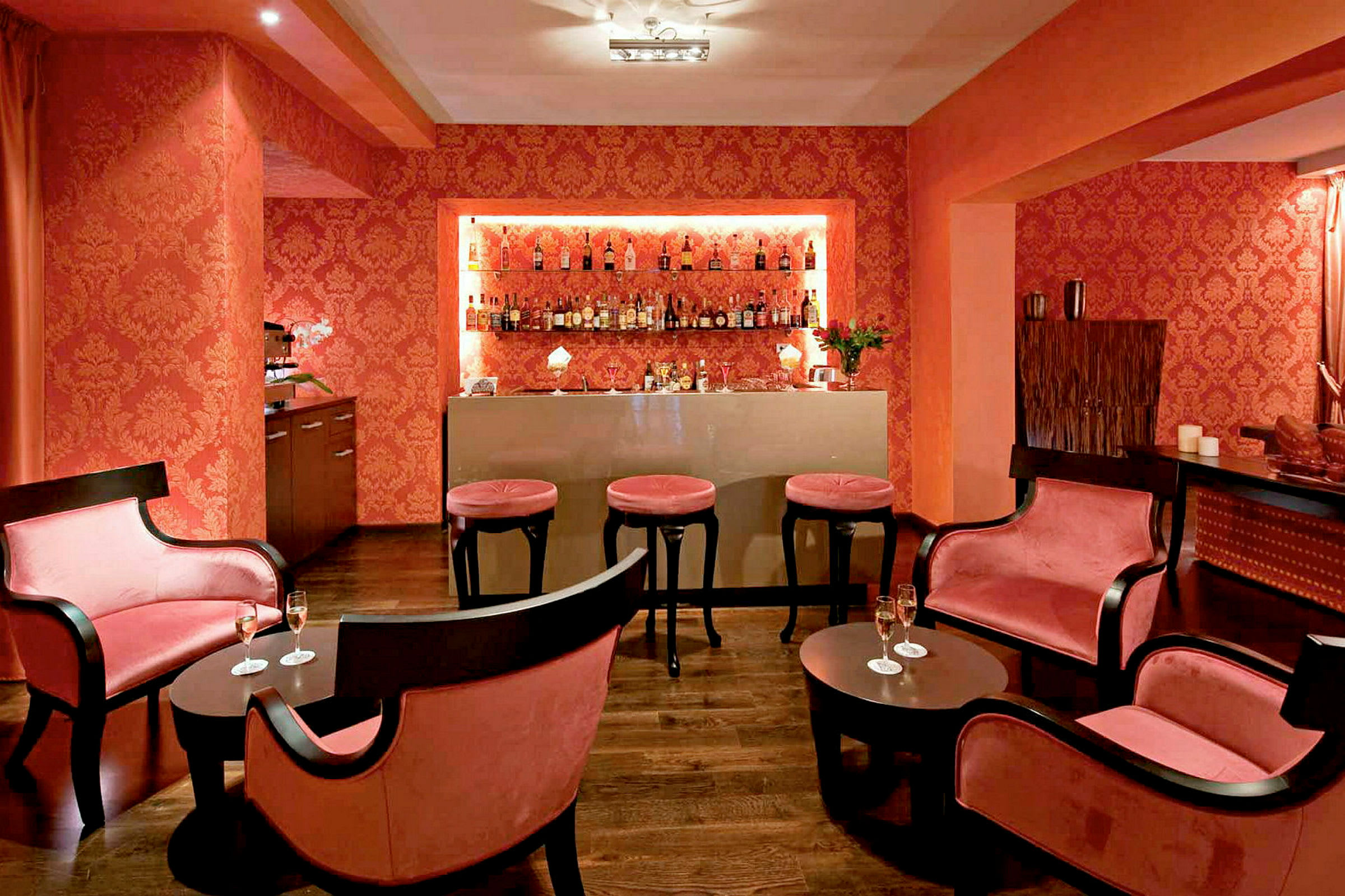 Billede der viser et elegant område indrettet som en cocktailbar med mørke møbler, lyserøde puder og flasker på en hylde. (AI-genereret tekst)