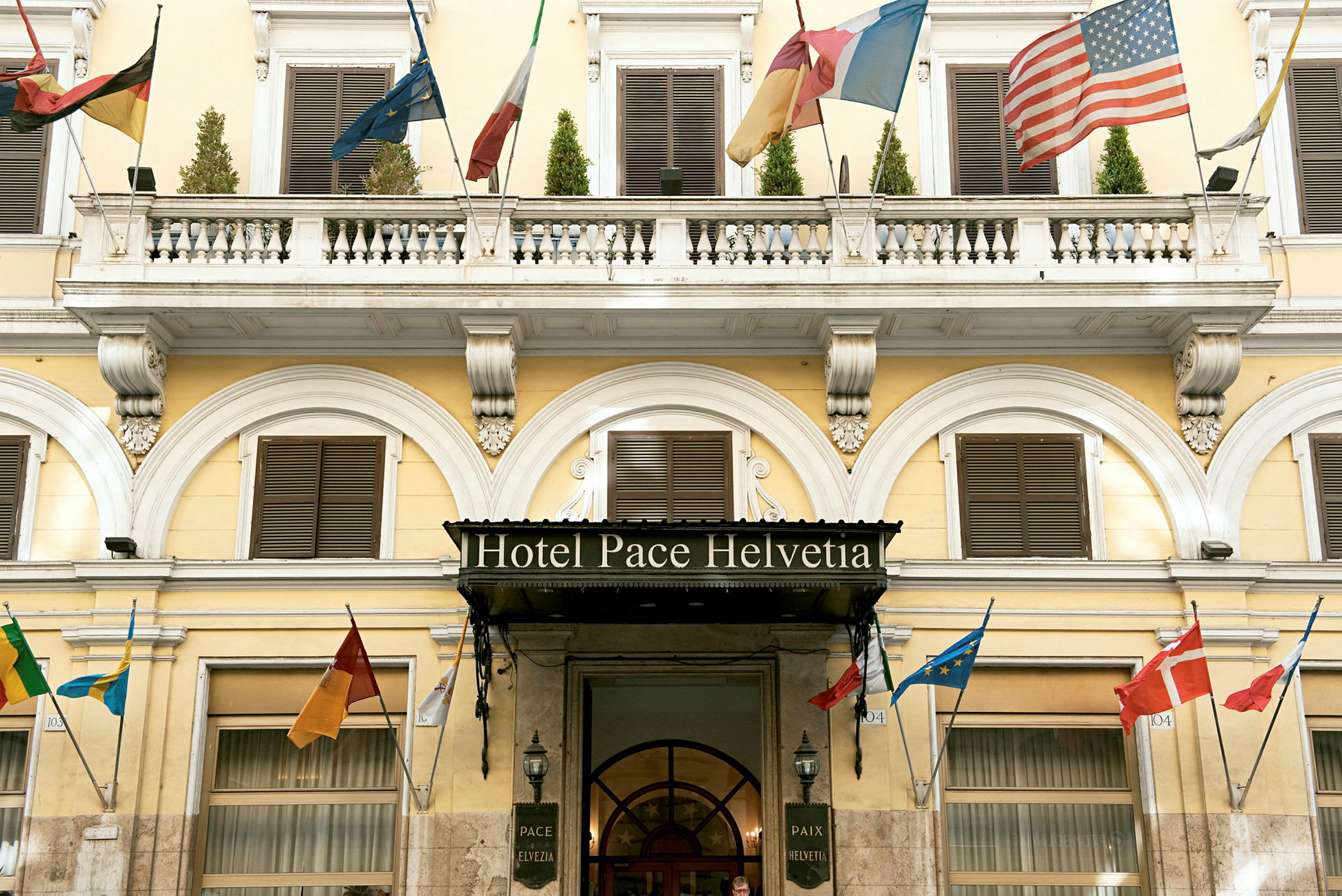 Hotelfacade med navnet 'Hotel Pace Helvetia', dekoreret med flere landes flag. (AI-genereret tekst)