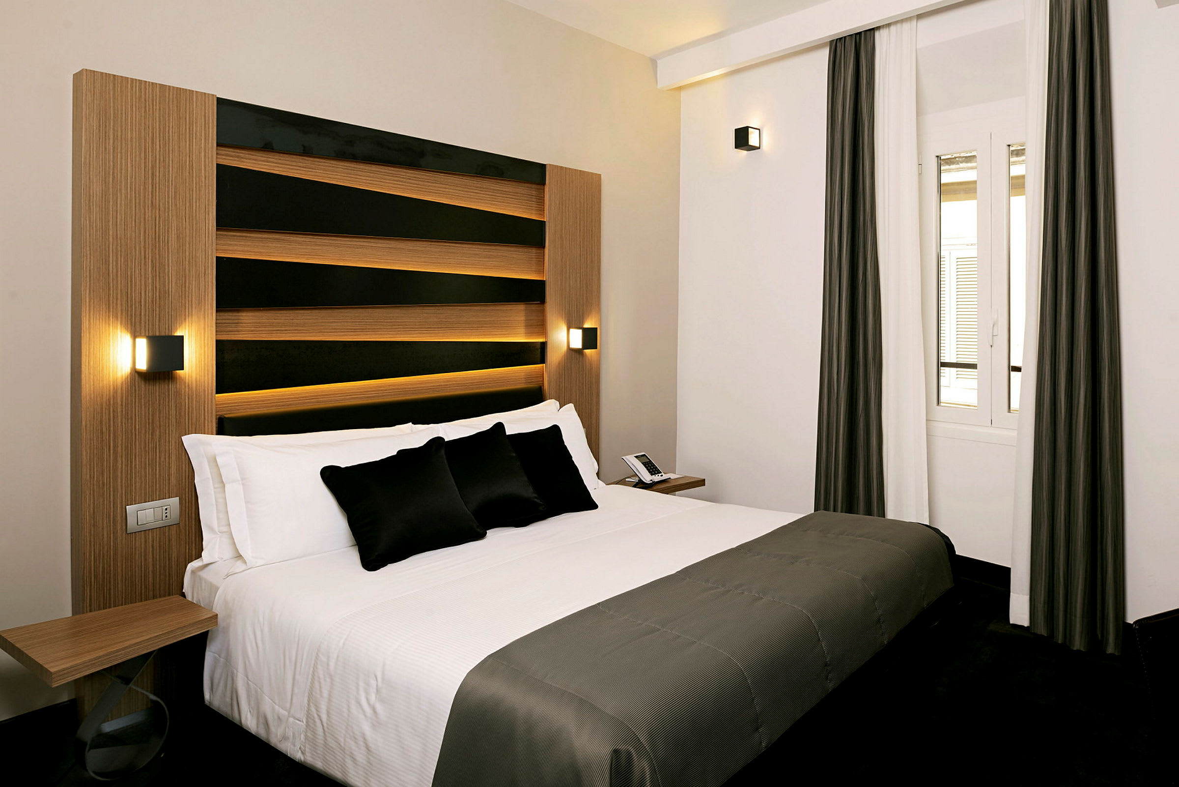 Dette billede viser et hotelværelse med en moderne og elegant seng med hvidt sengetøj og mørke dekorative puder. Værelset har en enkel og stilfuld indretning med et vindue med neutrale gardiner. (AI-genereret tekst)