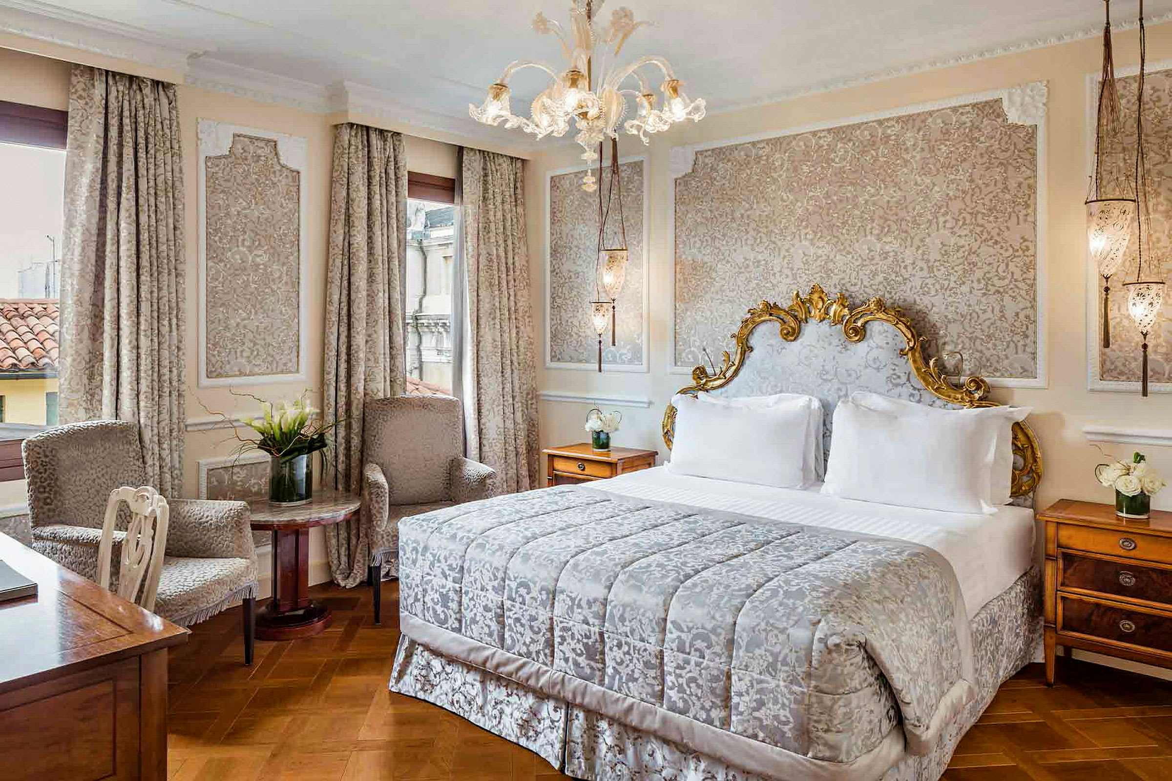 Et elegant soveværelse med en stor seng med dekoreret sengegavl, to lænestole og et bord med en vase med blomster. (AI-genereret tekst)