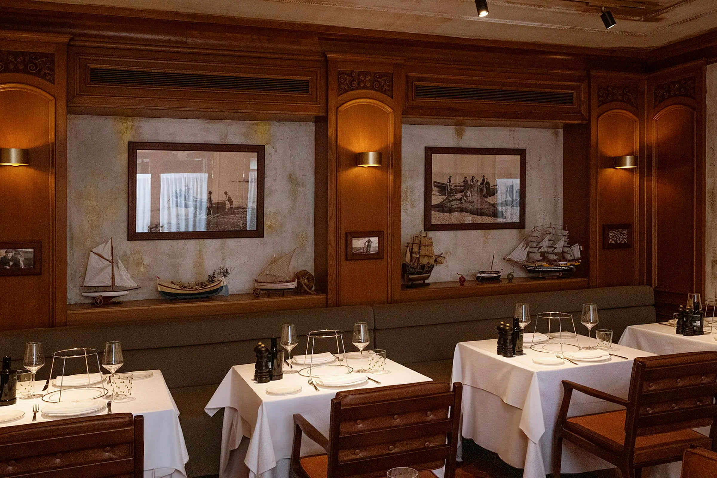 En elegant restaurantinteriør med borde dækket med hvide duge og træmøbler. Væggene er dekoreret med billeder og modelbåde. (AI-genereret tekst)