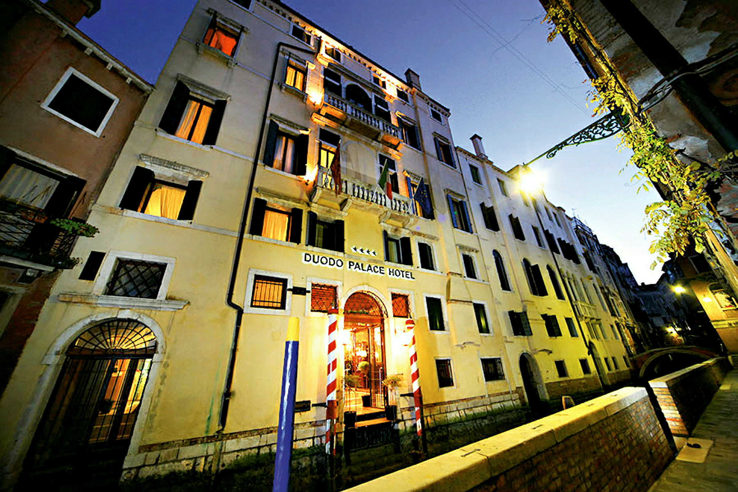 Lejlighedsbygning i tre etager med skilt Duodo Palace Hotel, omgivet af en kanal og en gangsti. (AI-genereret tekst)