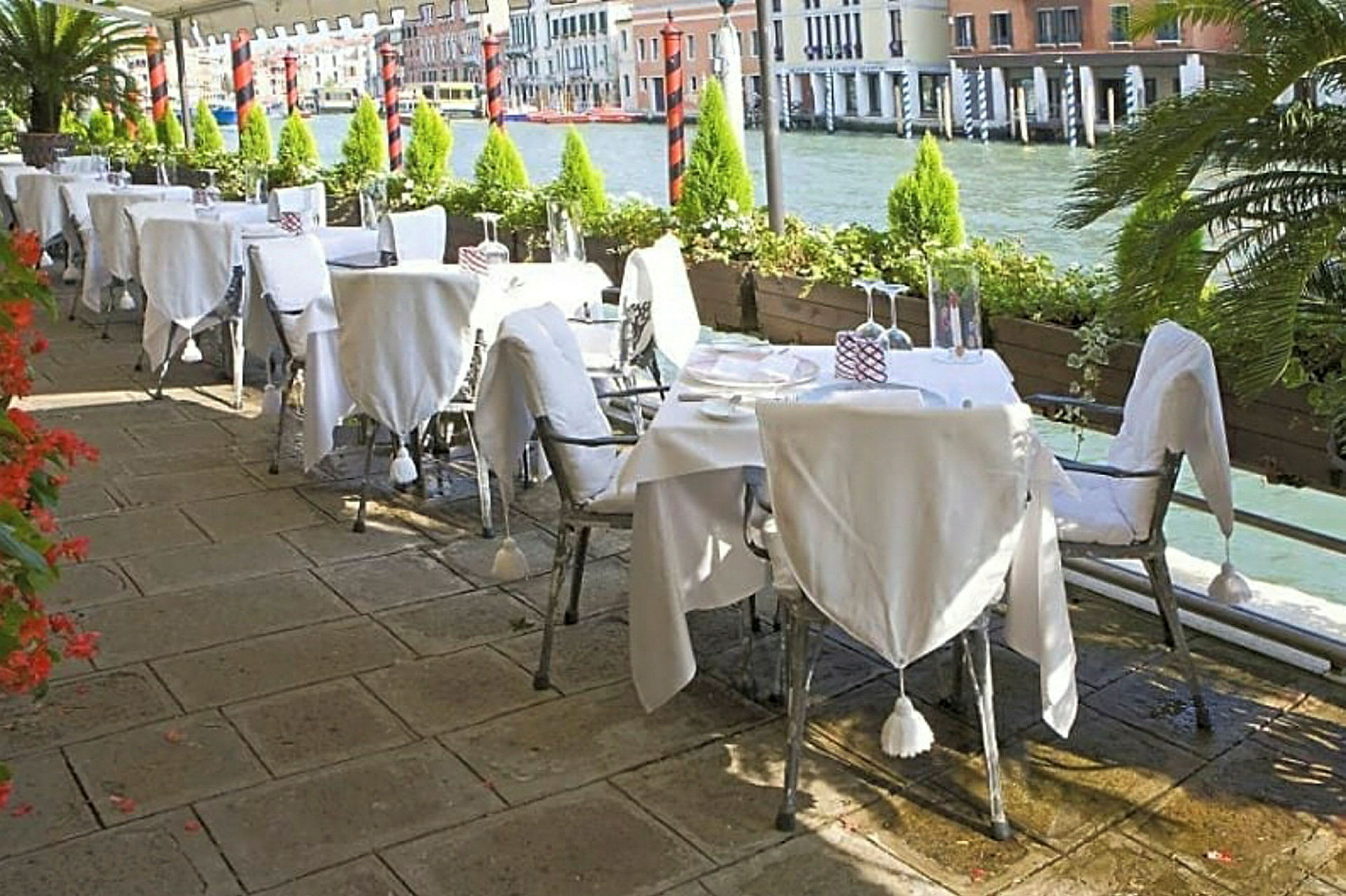 En udendørs restaurant med hviddugede borde ved en kanal med flere bygninger i baggrunden. (AI-genereret tekst)