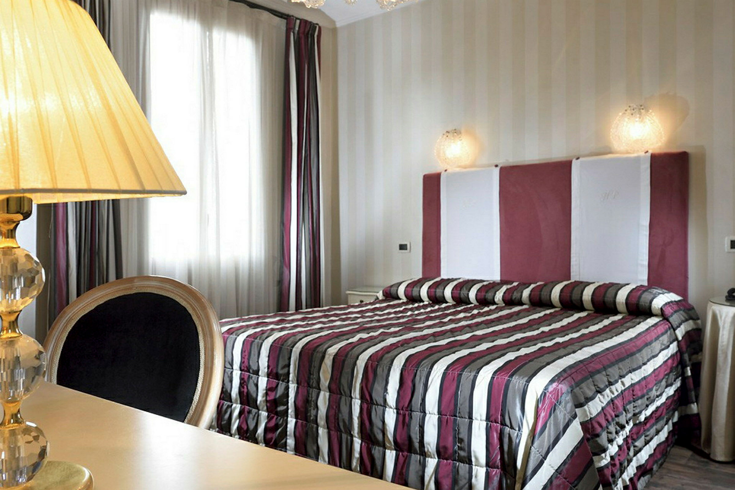 Et soveværelse med en seng med et stribet sengetæppe og en polstret sengegavl. Til venstre står en lampe på et bord. (AI-genereret tekst)