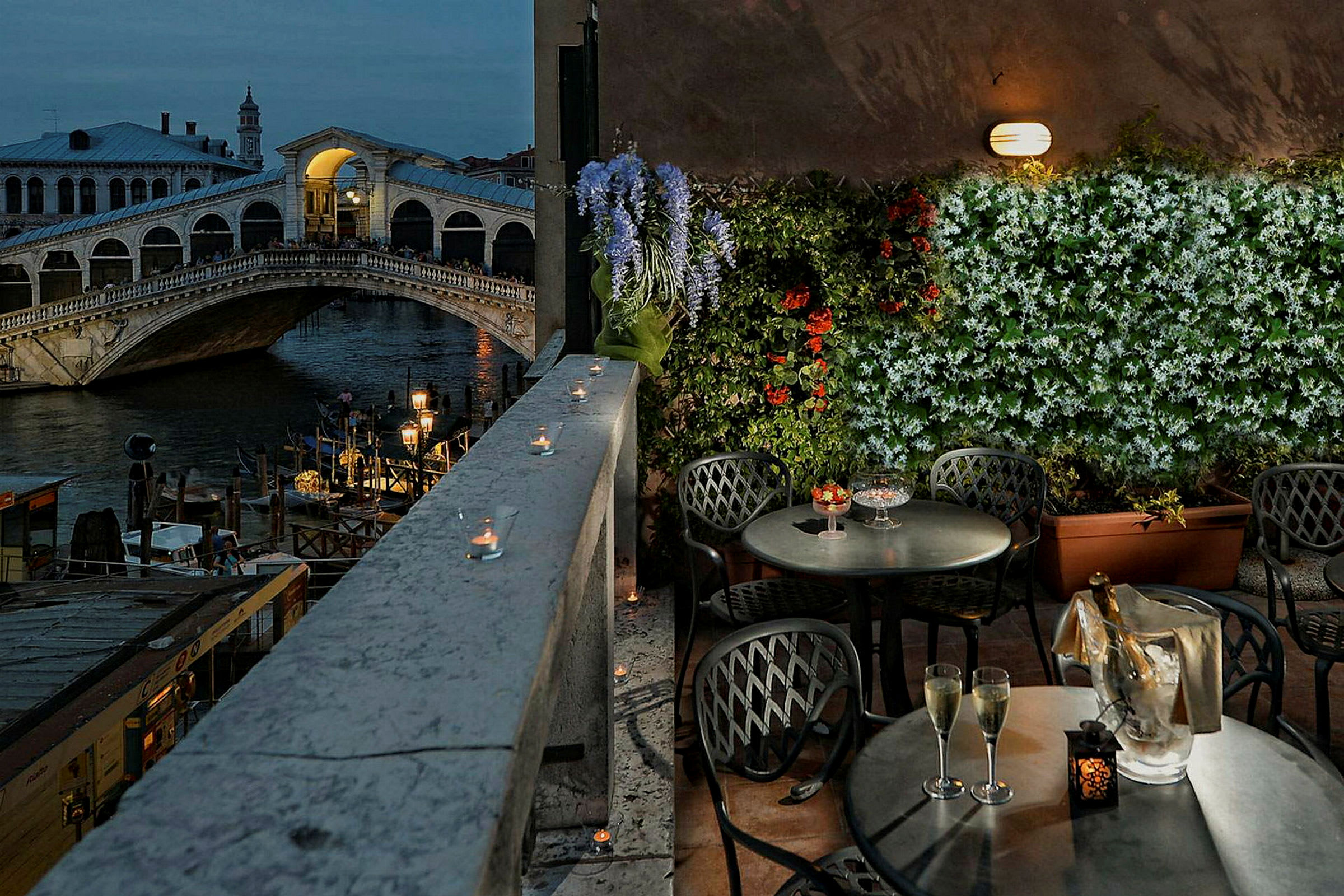 Billedet viser en terrasse med borde og stole, udsigt over Rialtobroen i Venedig, blomster og lys dekorerer området. (AI-genereret tekst)