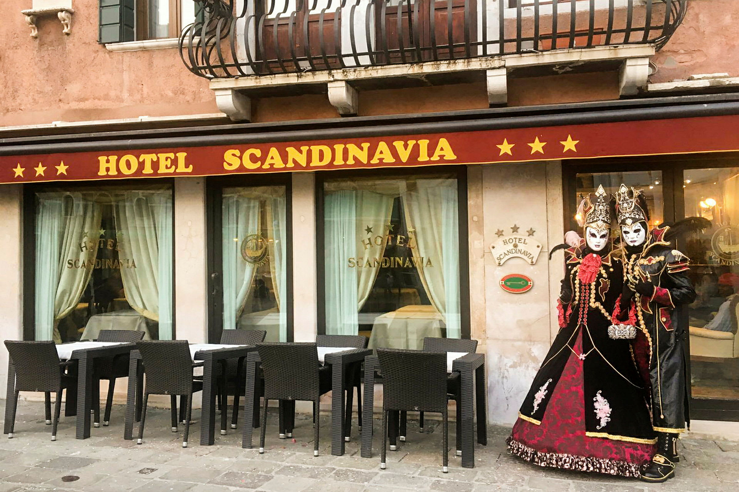 Hotellets façade med navnet 'Hotel Scandinavia', møblerede siddepladser udendørs og personer klædt i traditionelle maskeradedragter. (AI-genereret tekst)