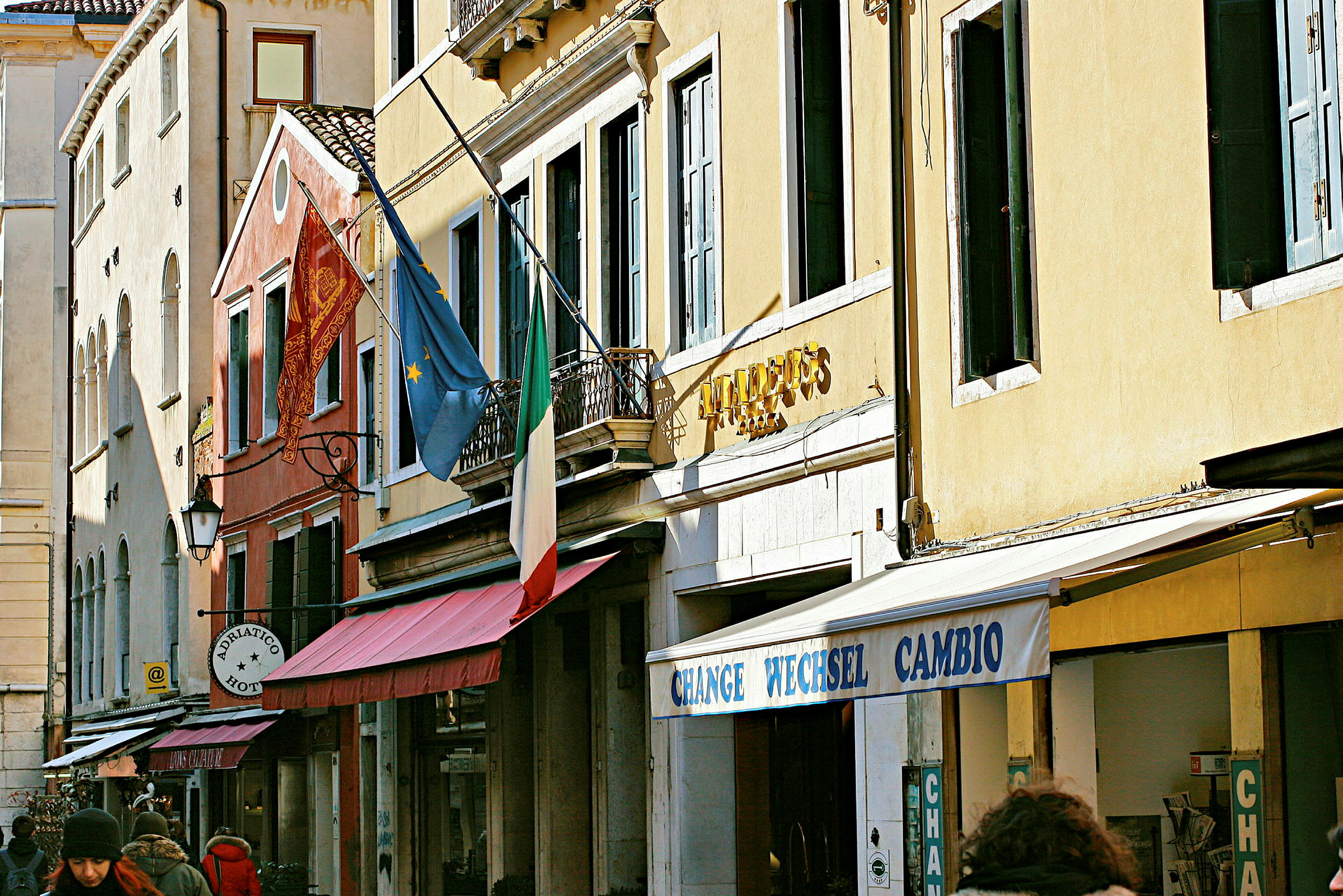 Facader af bygninger med et italiensk flag, europæisk flag og skilte, der angiver hotel og veksel. (AI-genereret tekst)