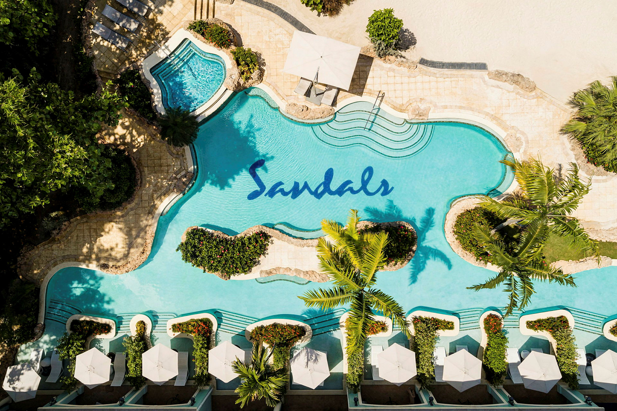 Et fugleperspektiv af en pool med Sandals-logoen, omgivet af planter og liggestole med parasoller. (AI-genereret tekst)