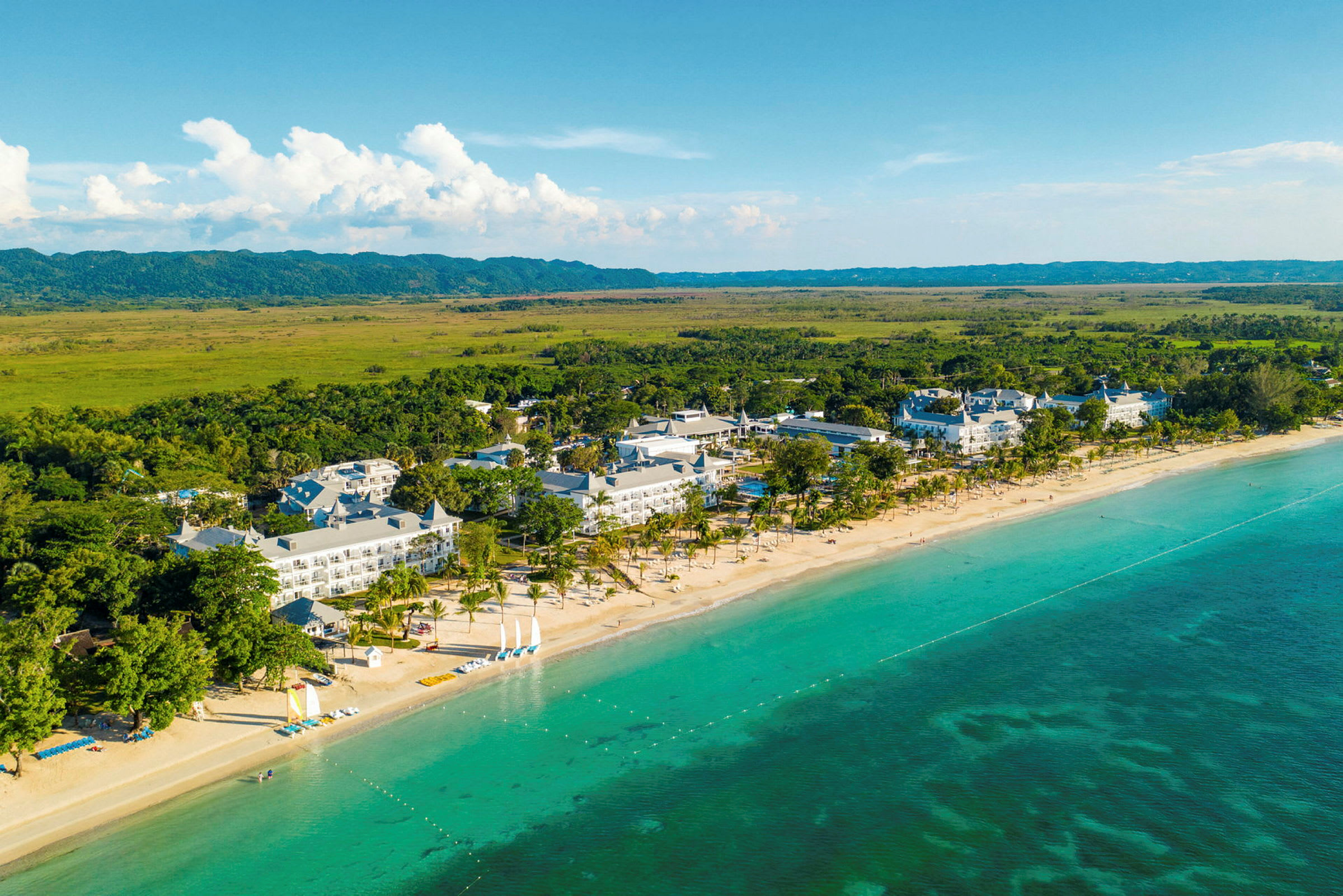 Luftfoto af et kystnært hotelområde med sandstrand, klart turkis havvand og omgivelser med vegetation. (AI-genereret tekst)