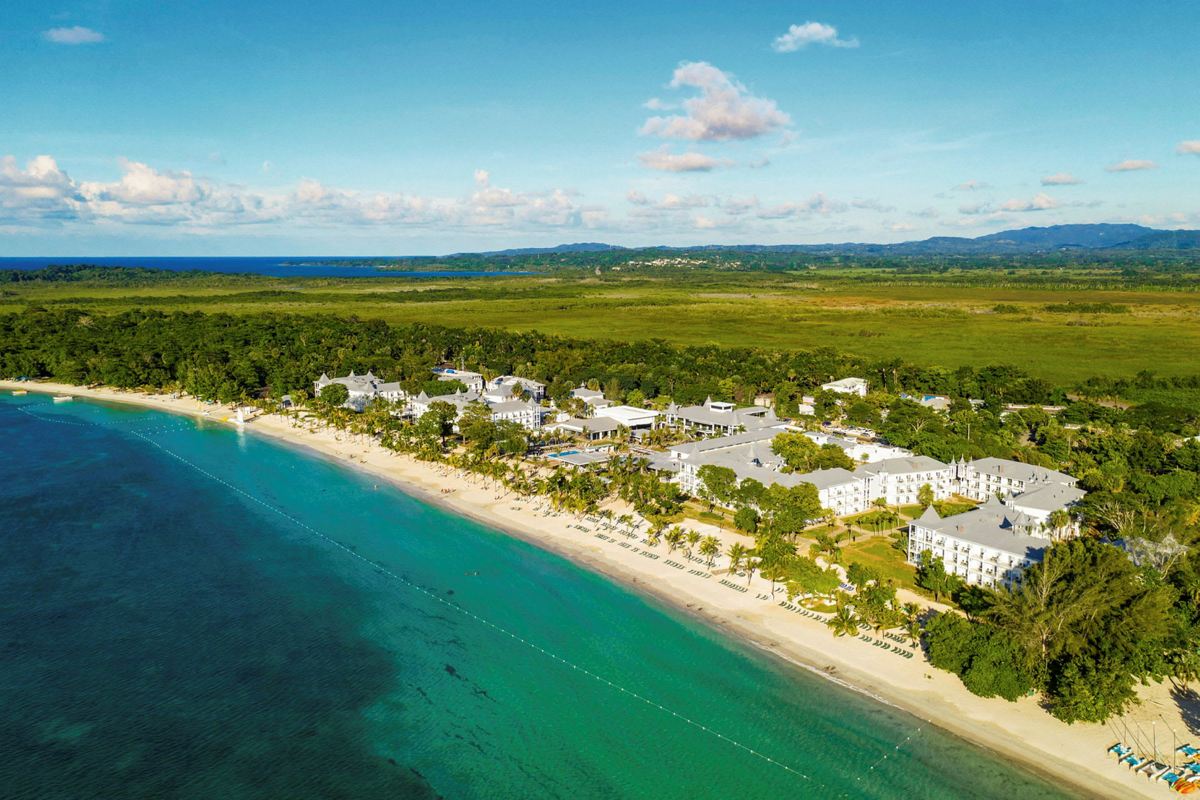 Luftfoto af en strand ved et turkisblåt hav og et hotelkompleks omgivet af frodig vegetation. (AI-genereret tekst)
