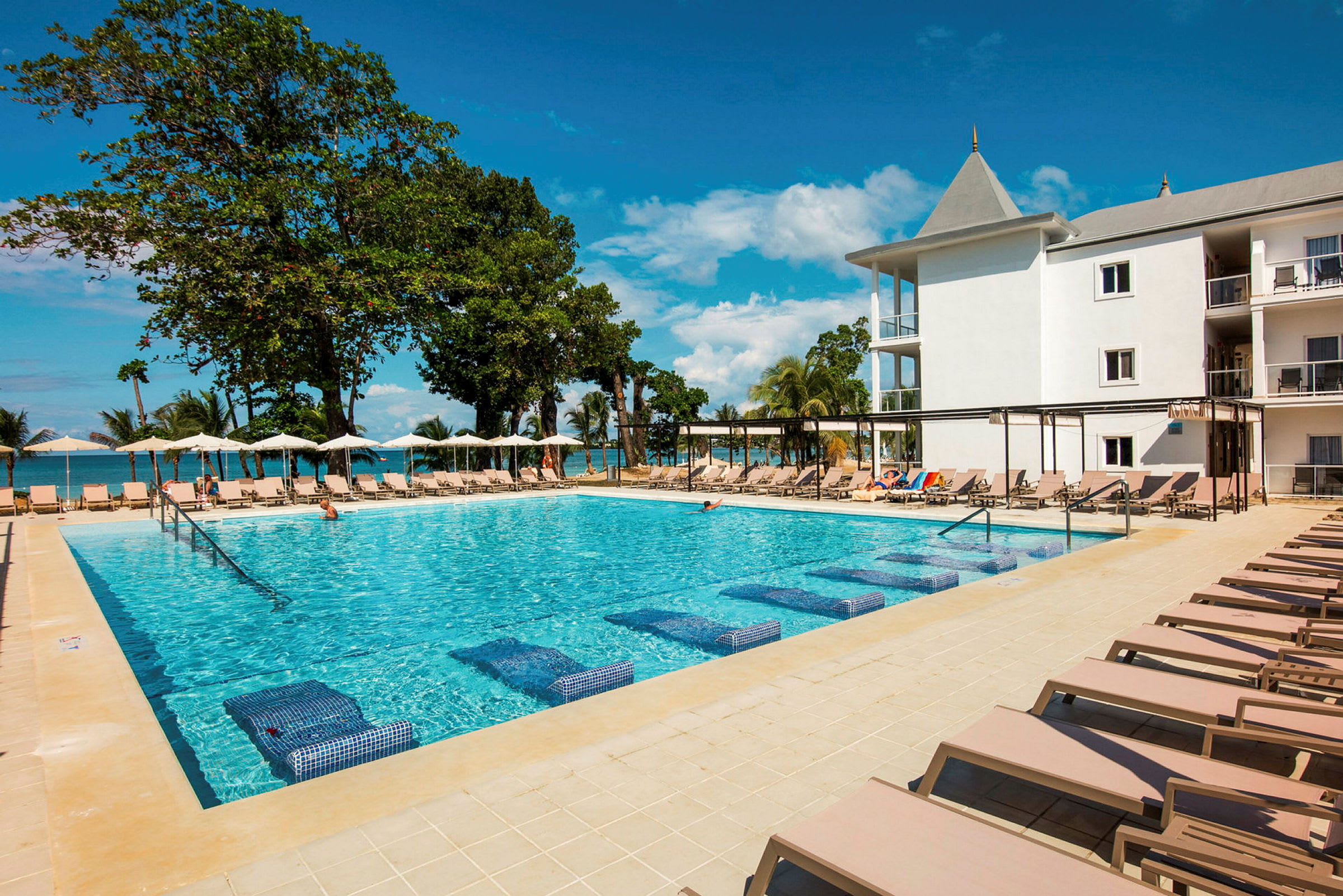 En krystalklar pool omgivet af solsenge med et hotel og træer i baggrunden. (AI-genereret tekst)