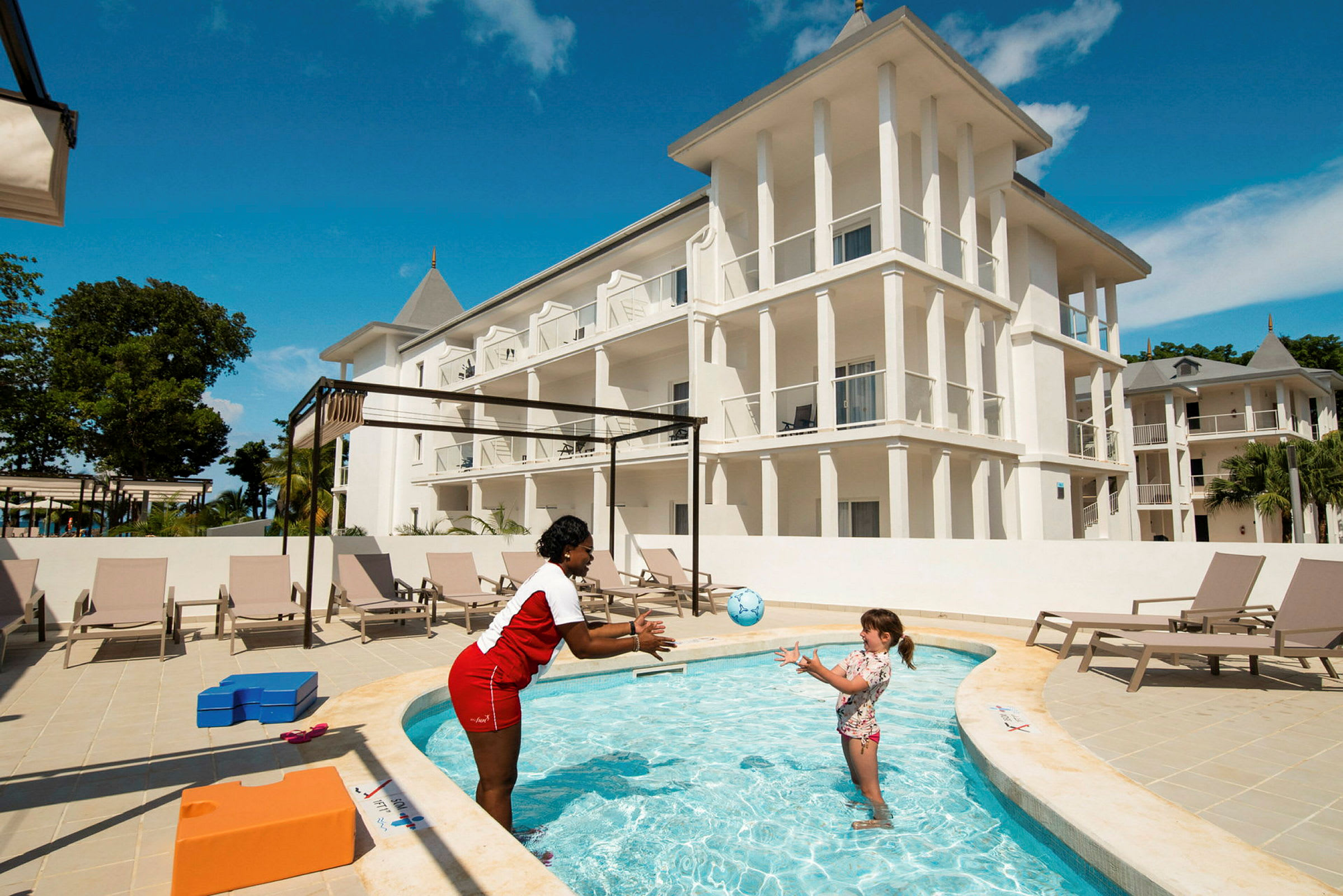 Et hotel med hvid facade og flere etager, foran en pool hvor en person kaster en bold til et barn. Liggestole er opstillet tæt på poolen. (AI-genereret tekst)