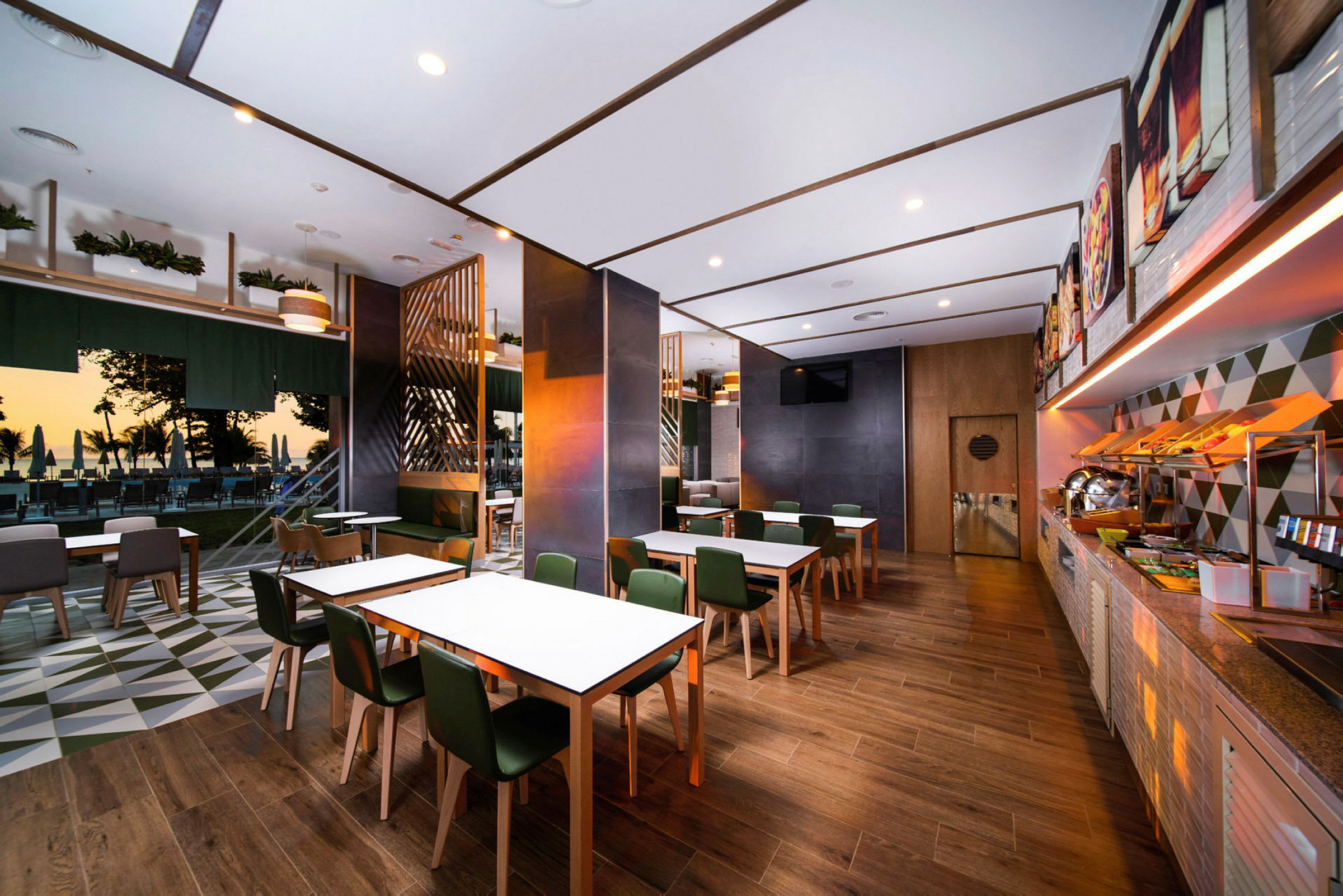 Et moderne restaurantområde med træmøbler, hvide borde og grønne stole. I baggrunden er der en udendørs serveringsplads med udsigt over stranden ved solnedgang. (AI-genereret tekst)