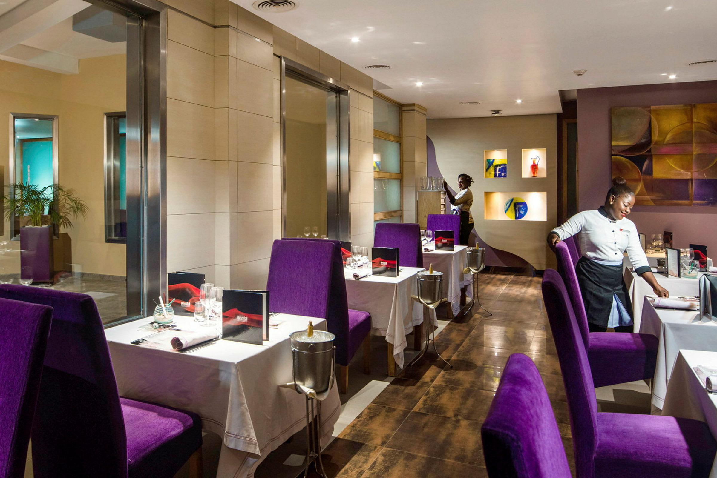 Billede af en elegant indrettet restaurant med lilla stole og hvide duge, samt tjenere der forbereder bordene. (AI-genereret tekst)