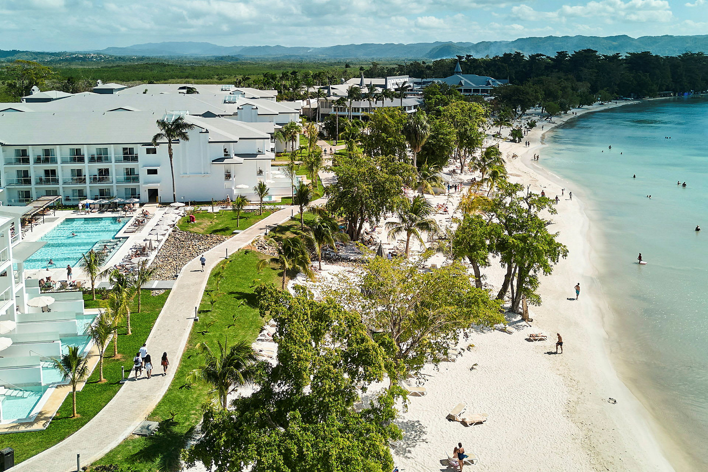 En sandstrand med personer, der svømmer i havet og hotelbygninger ved siden af en pool. (AI-genereret tekst)